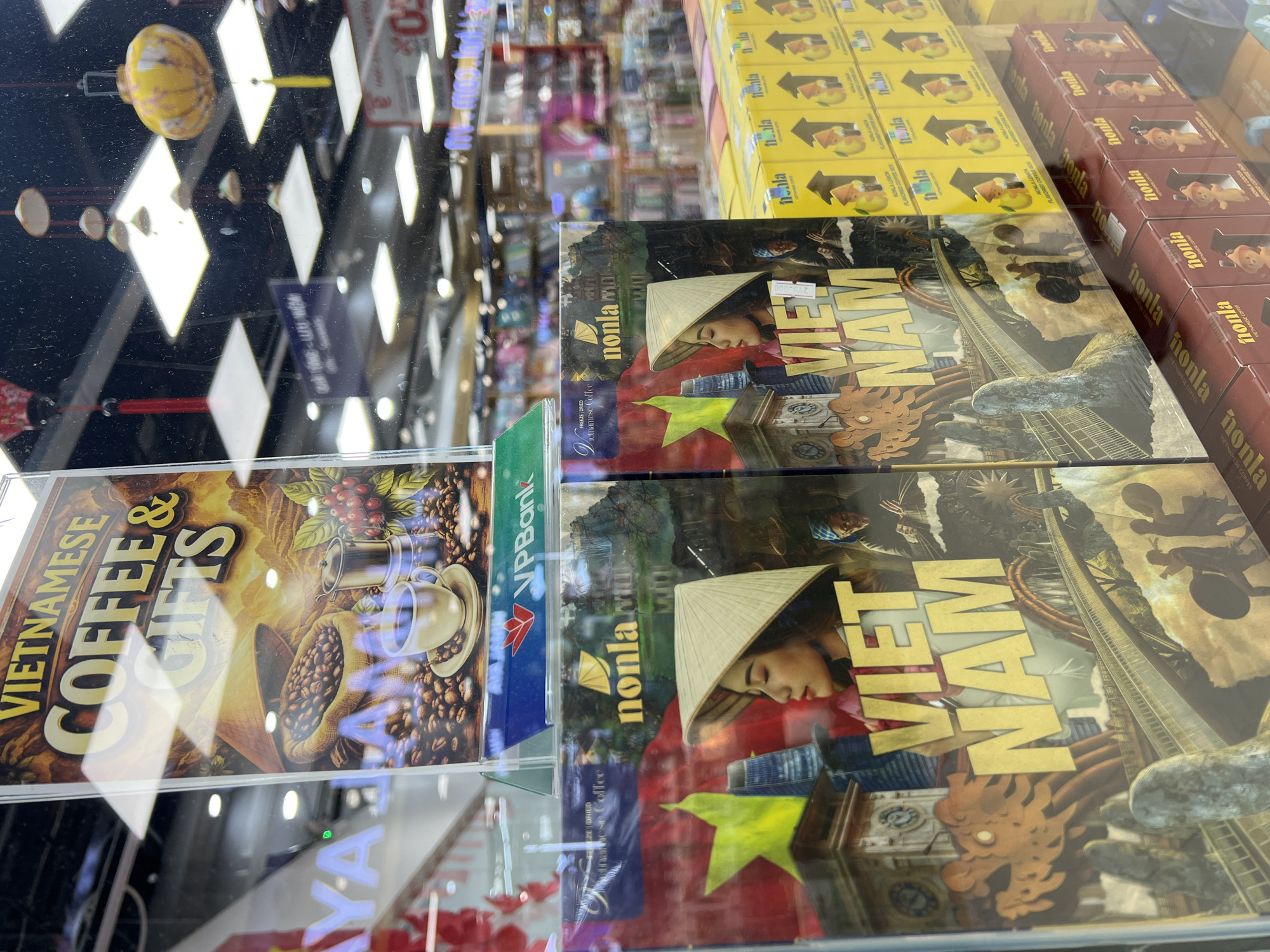 The image shows a display of Vietnamese coffee and gifts behind a glass surface, likely inside a store in Đà Nẵng, Vietnam. Featured items include boxes of Nonla brand freeze-dried Vietnamese coffee and flavored coffee, as well as gift boxes depicting Vietnamese national imagery, such as a woman wearing a conical hat and the Vietnamese flag. A sign advertises "Vietnamese Coffee & Gifts." Other items for sale are visible on shelves in the background.