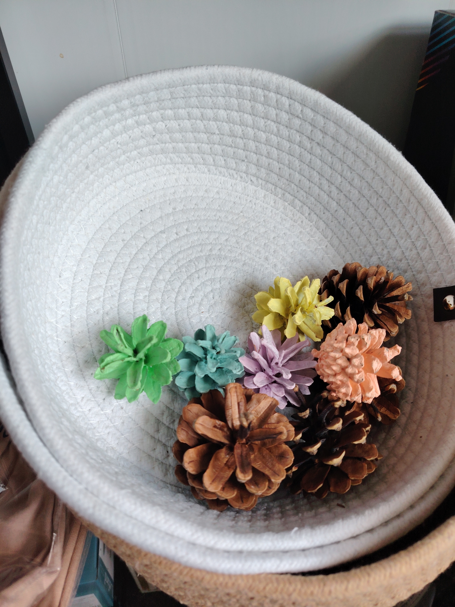 A high-angle shot shows a white woven rope basket with decorative pinecones. The pinecones are of various colors including brown, green, blue, yellow, purple, and orange. The basket is filled with these pinecones and rests on top of another larger woven basket. The background is a plain light-colored wall. The image does not depict any location in Konakovo, Russia or any recognizable urban elements.
