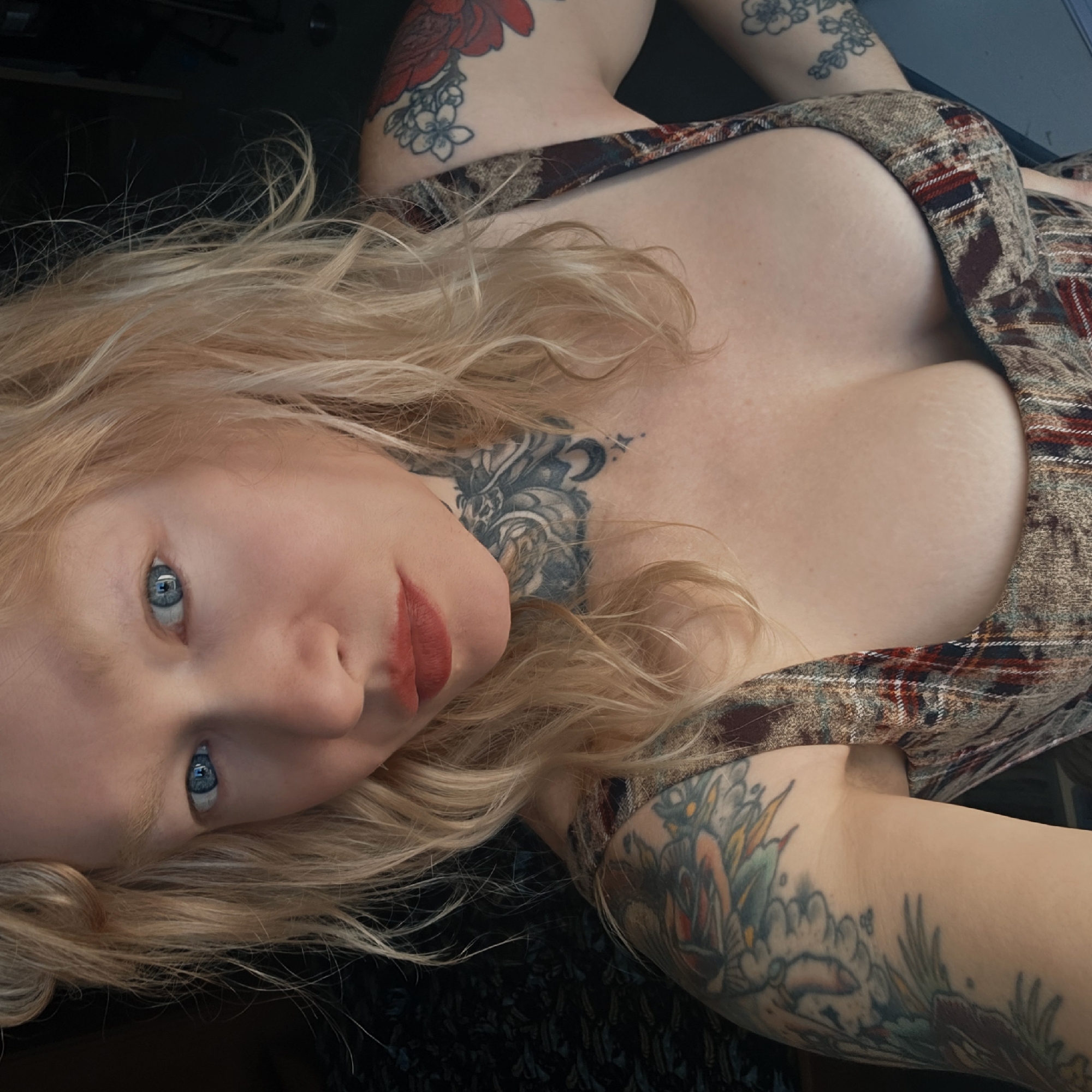 A woman with blonde hair, blue eyes, and a fair complexion is lying down, facing the camera. She has a tattoo on her neck and tattoos on both arms. She is wearing a plaid top. The background is dark. The location is not identifiable from the image as being in Saint Petersburg, Russia.