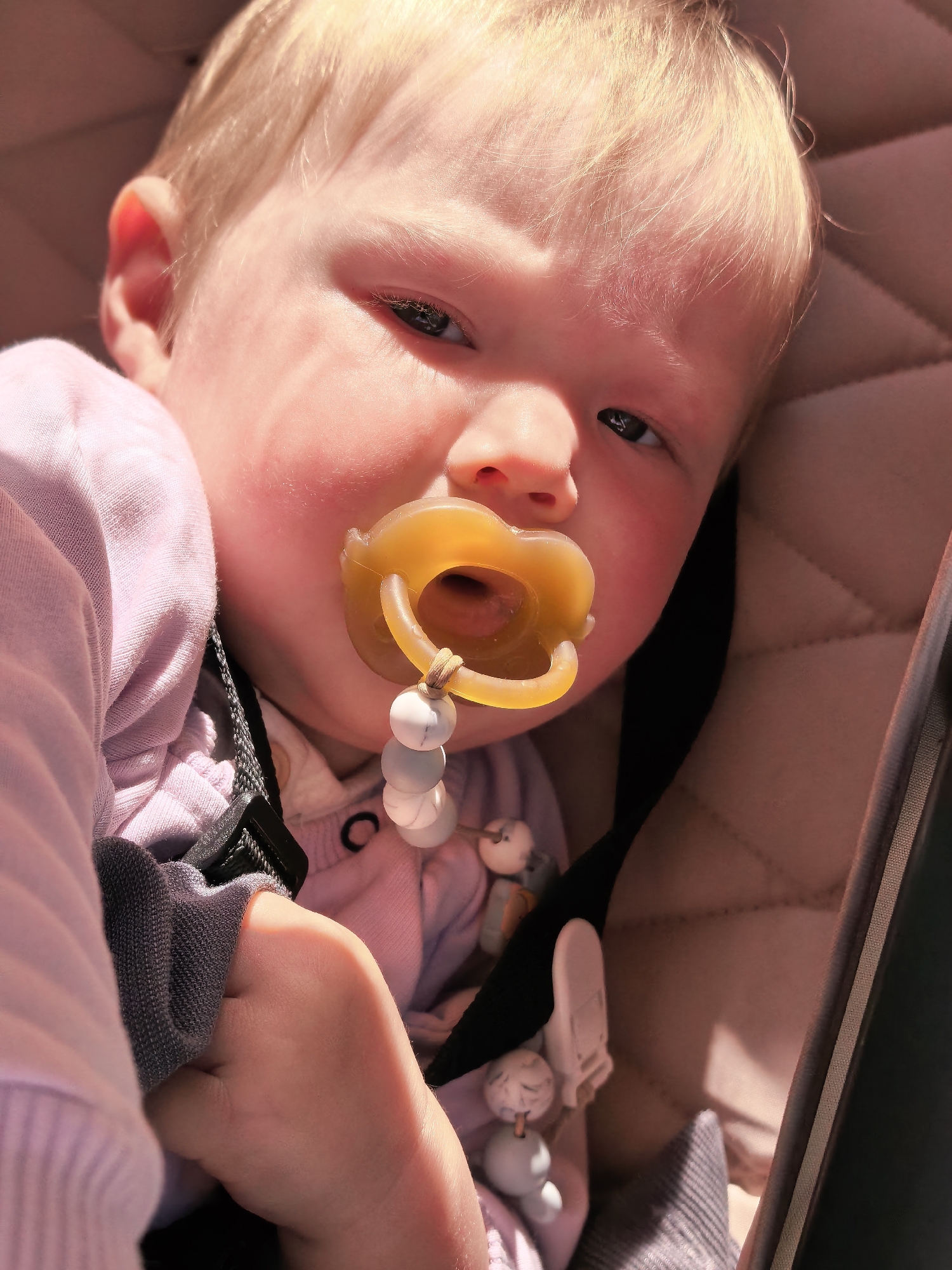 A close-up, eye-level shot shows a fair-skinned baby with blonde hair lying in a stroller. The baby has their mouth open, with a yellow pacifier in place and a beaded pacifier clip attached. The baby is wearing a pink long-sleeved shirt and a gray sleeve cuff is visible. The stroller's padded interior is a light brown color with a quilted pattern. The lighting suggests an outdoor setting with bright sunlight casting shadows. There is no indication of the city of Sterlitamak, Russia, in the image.