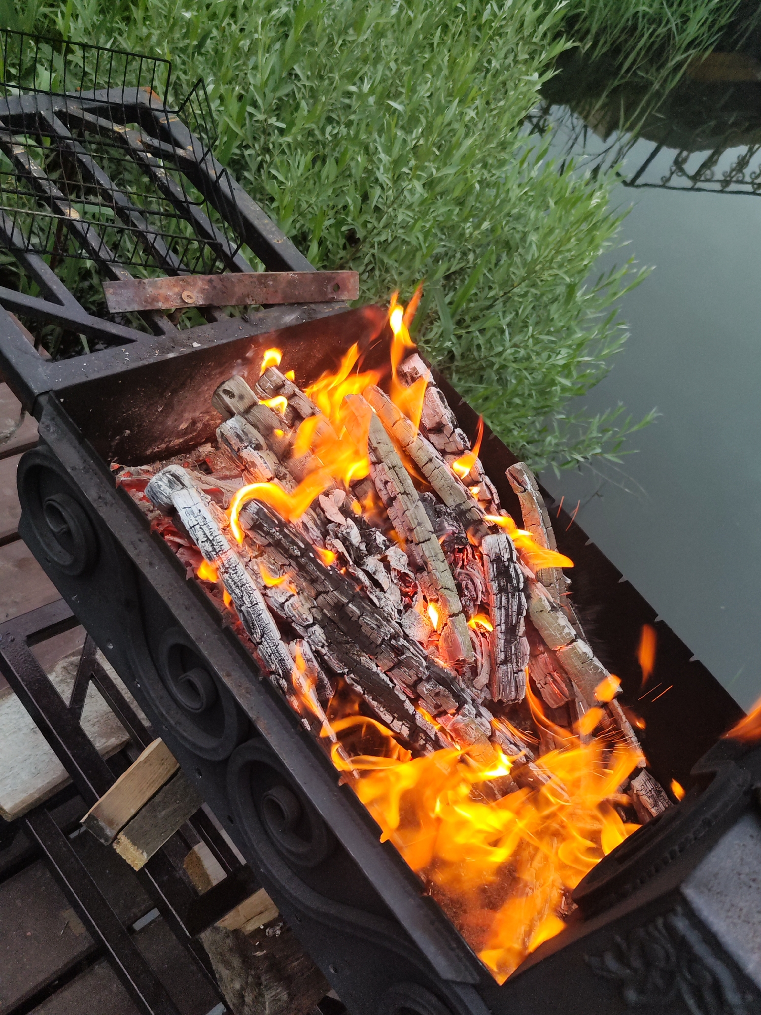A charcoal brazier filled with burning logs is positioned near a body of water. Adjacent to the water's edge, green vegetation is visible. The brazier appears to be made of dark metal and has decorative scrolling designs. In the upper left corner of the image, a metal grilling rack is partially visible. The scene suggests an outdoor setting, possibly for cooking or recreation. This image was taken in the city of Chaikovsky, Russia.