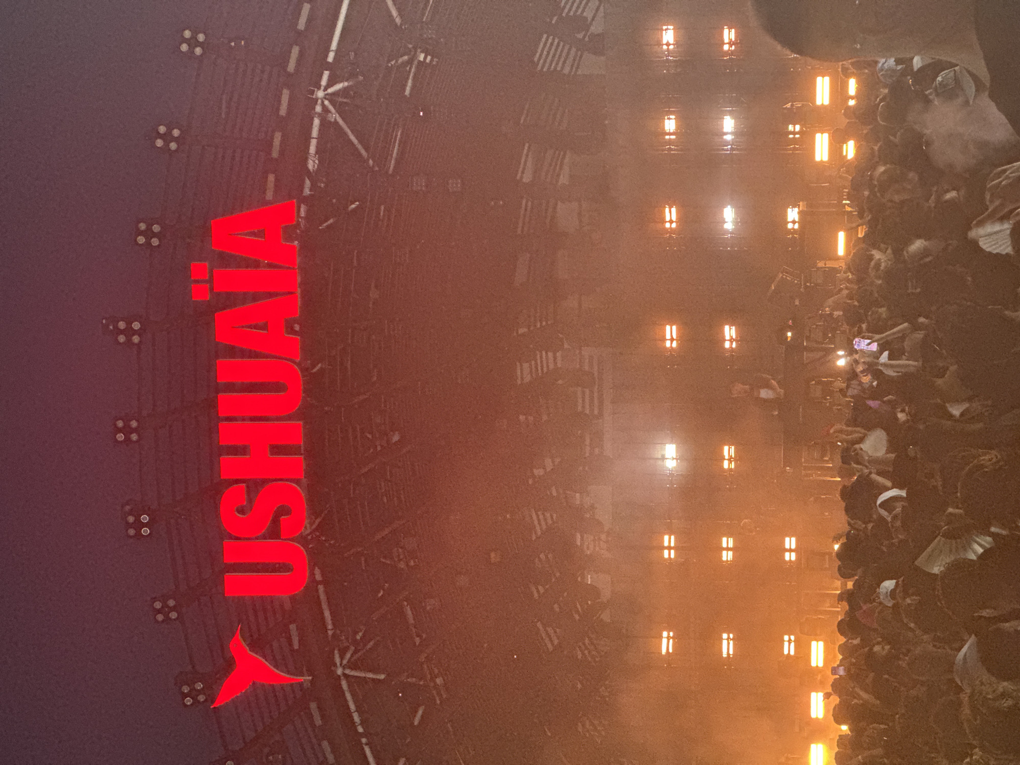 The image depicts a night-time outdoor concert or event at Ushuaïa Ibiza Beach Hotel in Playa d'en Bossa, Sant Josep de sa Talaia, Spain. A large crowd of people is gathered, facing a stage area where lighting and possibly a DJ setup are visible. Above the crowd, large red text spelling "USHUAÏA" with a stylized bird logo is illuminated, serving as a prominent branding element. The atmosphere appears to be energetic, with concert lighting creating a dramatic and hazy effect. The background shows the venue's infrastructure, including lighting rigs and structural elements.