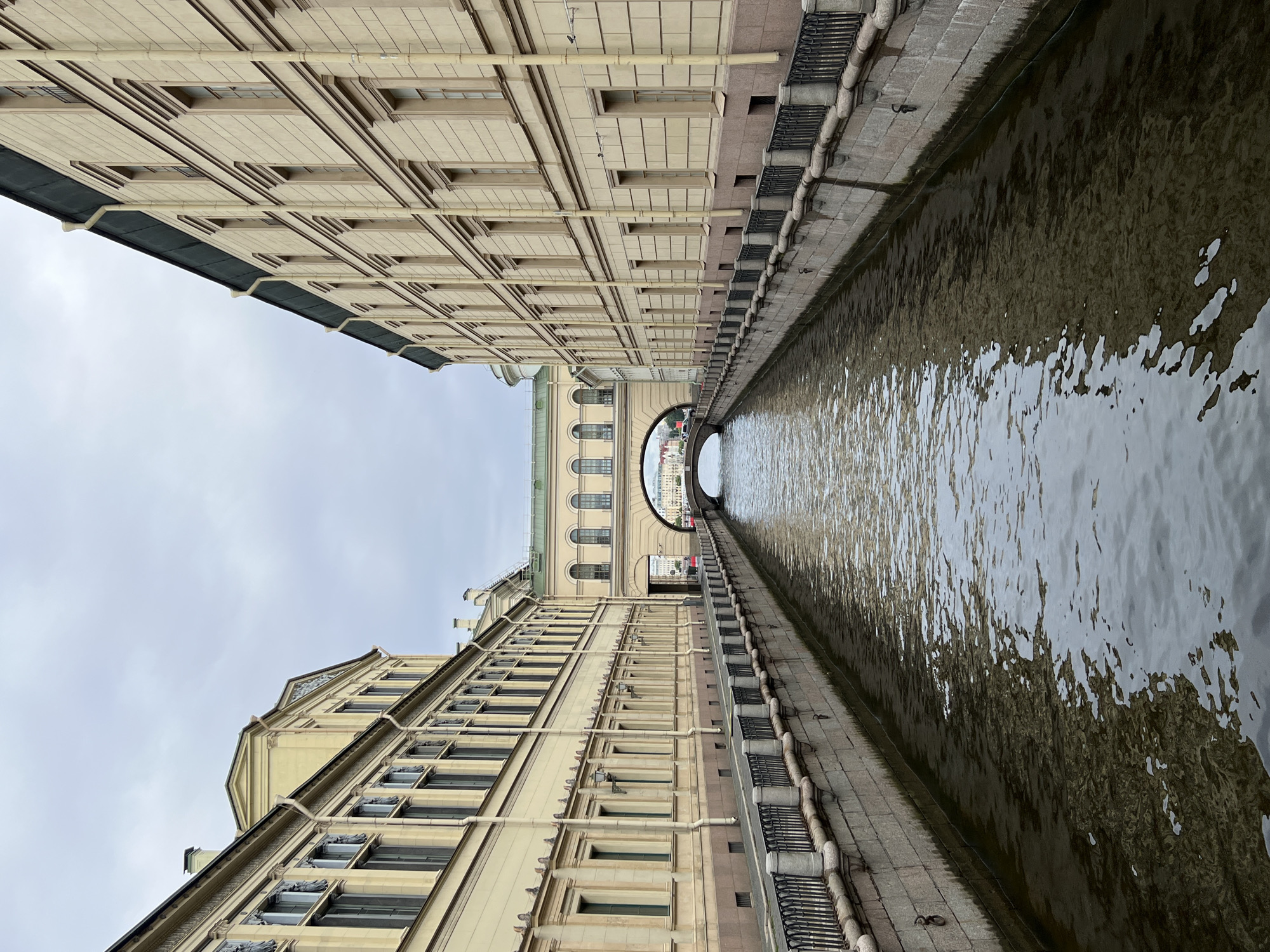 The image shows a canal in Saint Petersburg, Russia, bordered by tall, elegant buildings on both sides. The architecture is classical, with rows of windows and decorative elements. A distinctive arched bridge spans the canal in the distance, connecting the buildings above the water. The sky is overcast, casting a muted light on the scene. The canal's water is dark and reflects the buildings and the sky, with ripples indicating gentle movement. A dark metal railing runs along the stone embankment of the canal. No people are prominently visible, suggesting a quiet moment. The overall impression is one of historic urban beauty and serene canal-side architecture.