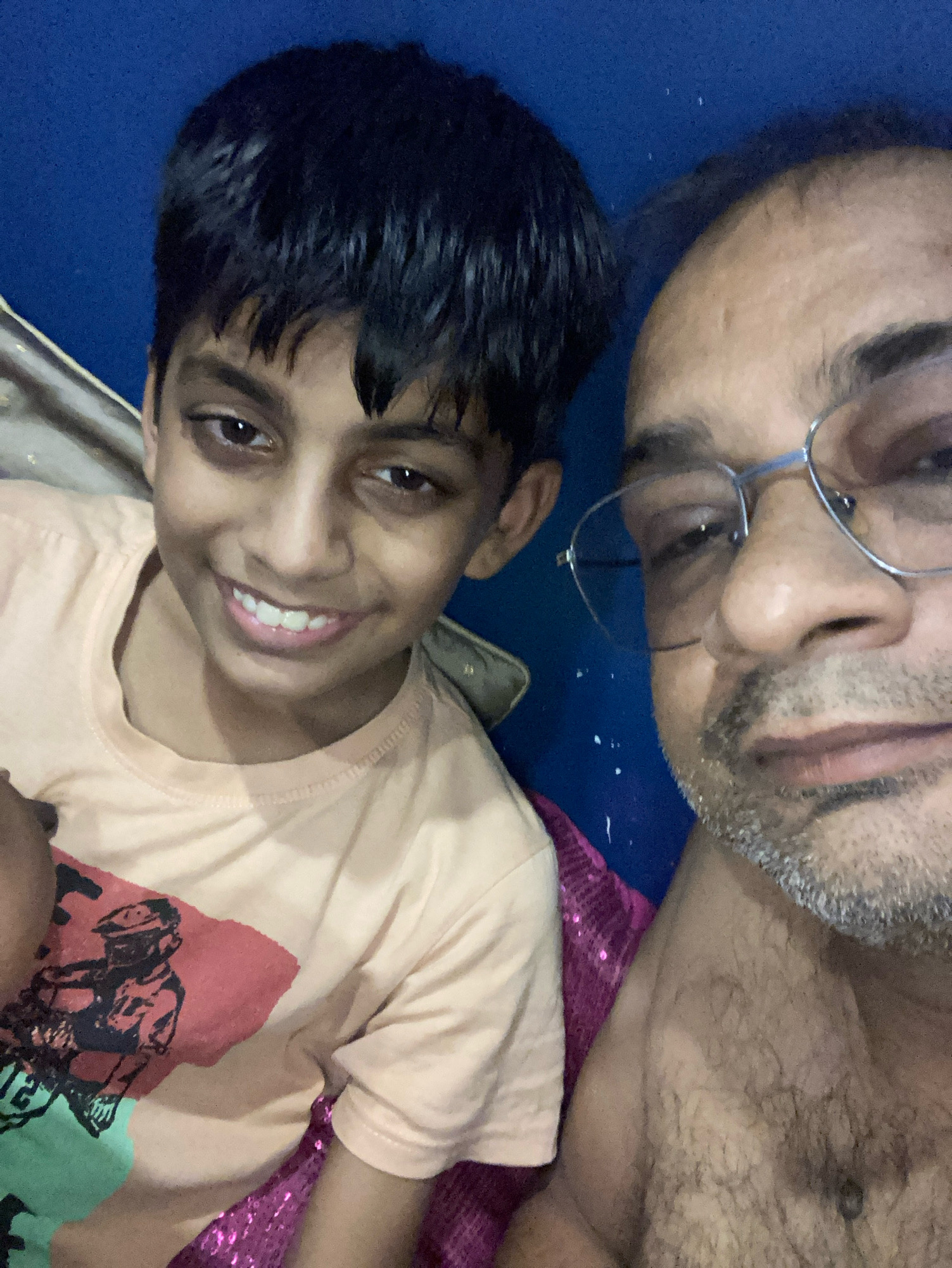 This is a medium shot selfie of two people, likely a father and son, with the camera positioned close to their faces. The background is a solid blue wall. The subject on the left is a young boy with dark, short hair and a wide smile, showing his teeth. He is wearing a light peach t-shirt with a graphic design featuring a red shape and a figure that appears to be riding a vehicle, with some green elements. The subject on the right is an adult male with glasses, a light beard, and a bald head with some hair on the sides. He is looking towards the camera. The context provided indicates the location is Kolkata, India. The lighting suggests it is indoors and possibly evening or night. The overall emotion conveyed is one of warmth and connection between the two individuals. There is no visible text in the image.