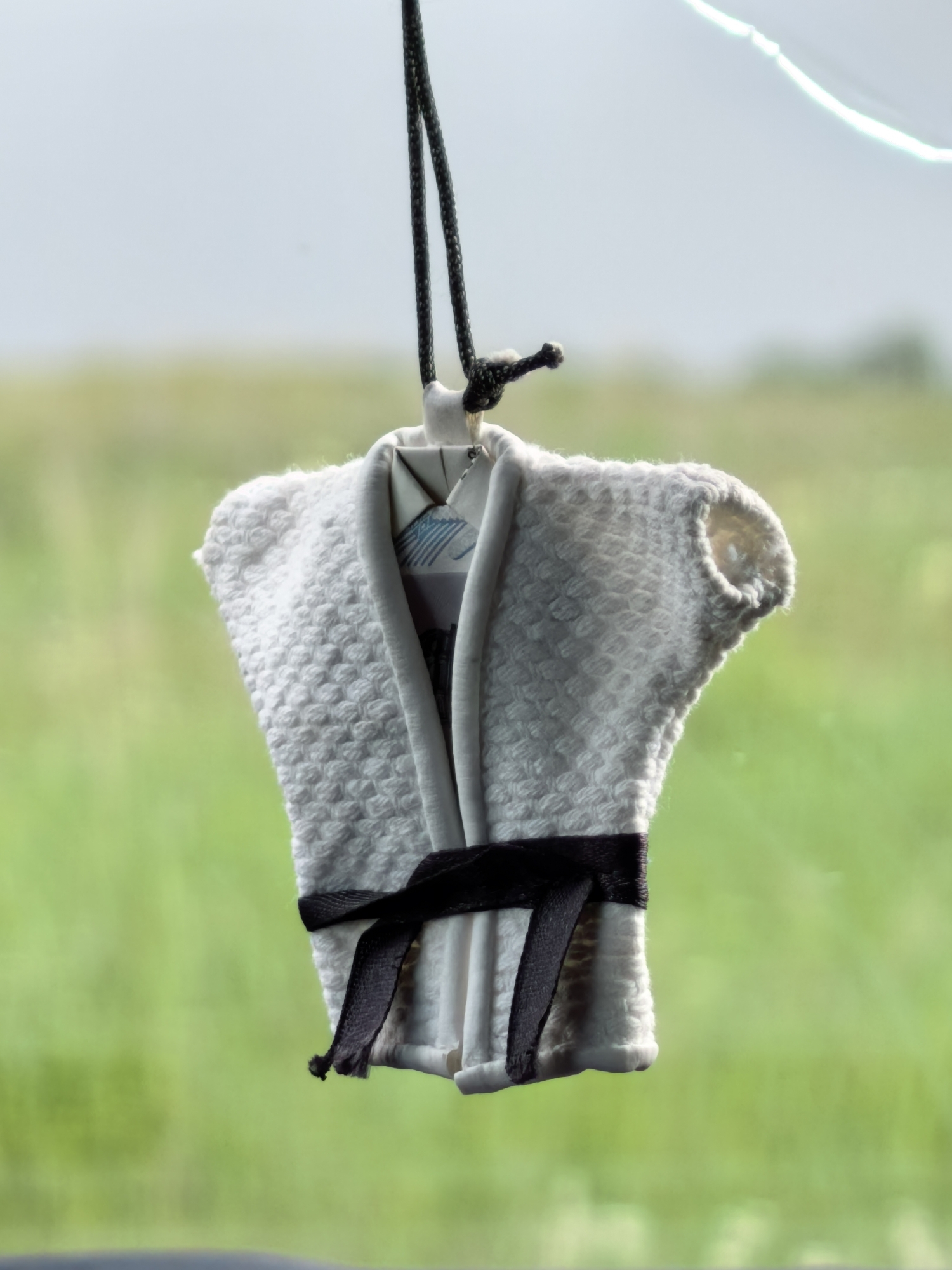 A miniature white karate gi, complete with a black belt, hangs from a black cord against a blurred background of green foliage and a pale sky. The gi is intricately textured, mimicking the weave of a real uniform. A small, patterned piece of paper is visible inside the collar. The scene appears to be captured through a window, possibly in a car, given the slight distortion and the hint of a dark interior at the bottom of the frame. There are no people or other discernible objects in the immediate vicinity. The lighting suggests an outdoor, daytime setting. There is no information to suggest this location is in the city of Кузьминовка, Russia.