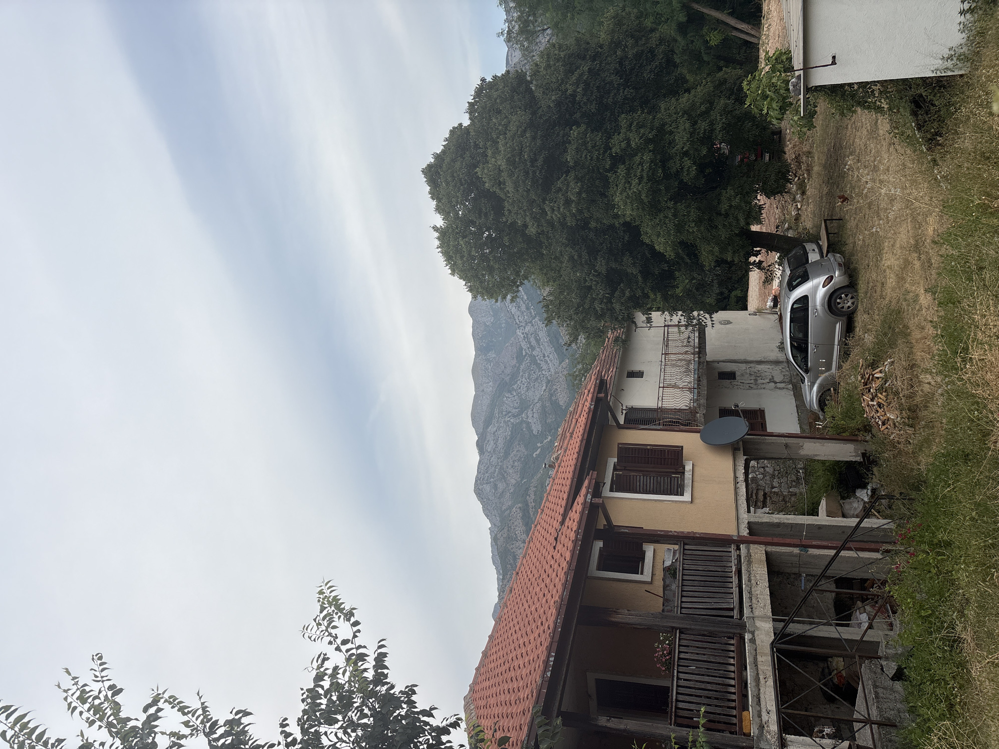 The image depicts a scene in Stari Bar, Montenegro, with a focus on a residential building and its surroundings. In the foreground, a portion of a multi-story building with a red-tiled roof is visible, featuring wooden shutters on its windows and a balcony with a weathered wooden railing. To the right of this building, a silver car is parked on a grassy, unpaved area. Behind the buildings, a large, dense green tree dominates the mid-ground, with a backdrop of rugged, grey mountains under a hazy, overcast sky. The overall setting appears to be a somewhat rural or semi-developed area, with a mix of natural and man-made elements. The light suggests it is daytime, likely overcast, with no clear indication of a specific time. There is no visible text in the image.