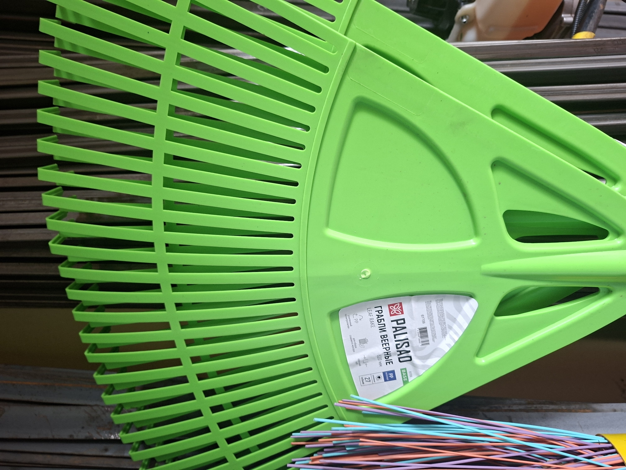A close-up, overhead shot of a bright green plastic leaf rake is featured, taking up most of the frame. The rake's tines, which are numerous and closely spaced, extend upwards and outwards, fanning out from a central point. The rake's body has a textured, molded surface with visible internal ribbing for support.
To the left of the rake, a bundle of thin, colored plastic rods, likely broom bristles or similar material, protrude from a yellow container, adding a vibrant contrast to the green rake. These rods are an assortment of pink, purple, blue, and orange.
On the surface of the rake, a white label is affixed. The label contains text in both Cyrillic and Latin alphabets. The Cyrillic text reads "ГРАБЛИ ВЕЕРНЫЕ" (Granuly Veer'nyye), which translates to "Fan Rake." Below this, "PALISAD" is prominently displayed in large, bold Latin letters, along with other product information including model numbers and material composition (PP).
The background is composed of a dark, metallic, slatted surface, possibly a fence or storage unit, which is out of focus. In the far right background, a portion of an orange plastic container, possibly part of a garden tool or machine, is visible.
The overall impression is a product shot, highlighting the design and details of the leaf rake. The context of Шклоў, Belarus, is not directly evident from the image itself, but the presence of Cyrillic text on the product label suggests a connection to that region.