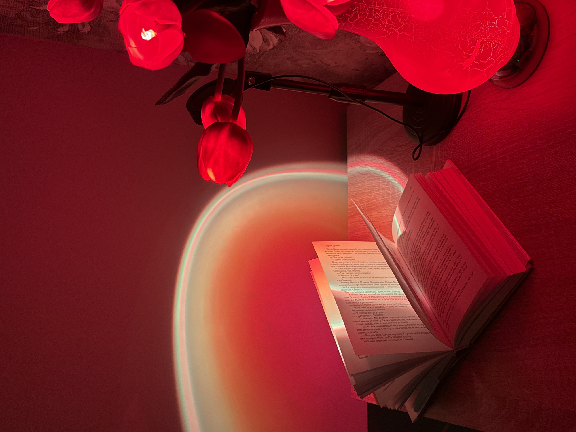The image depicts an indoor scene illuminated by a red sunset lamp. In the foreground, an open book lies on a wooden surface. To the right, a vase holds several red tulip-like artificial flowers, some of which are lit by the lamp. Behind the flowers, a black lamp with a flexible neck is visible. The background features a wall bathed in the projected red and orange light from the sunset lamp, creating a gradient effect. The scene is devoid of any people or recognizable urban or natural landmarks.