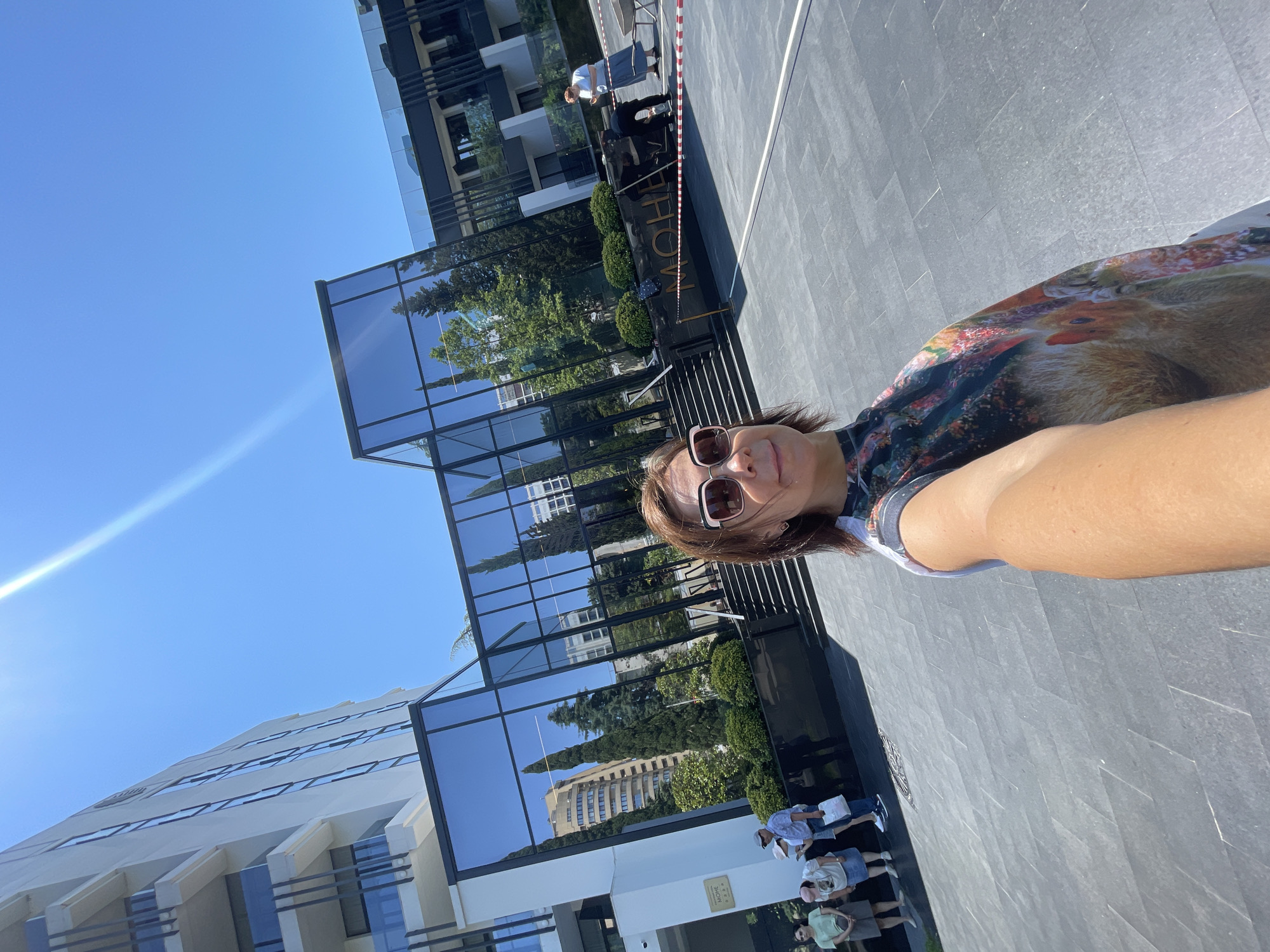 A woman is taking a selfie outdoors in front of a modern building with a glass facade. The building appears to be a hotel or resort, with lush landscaping and trees surrounding the entrance. The woman is wearing sunglasses and a colorful dress with a print of what appears to be animal faces. In the background, several other people are visible, some standing and looking towards the building. There is also caution tape across the entrance, suggesting some form of closure or construction. The sky is clear and blue, indicating a sunny day. The location appears to be Sochi, Russia, as evidenced by the signage on the building which includes Cyrillic text, specifically the word "MORE." The architecture and landscaping suggest a high-end establishment.