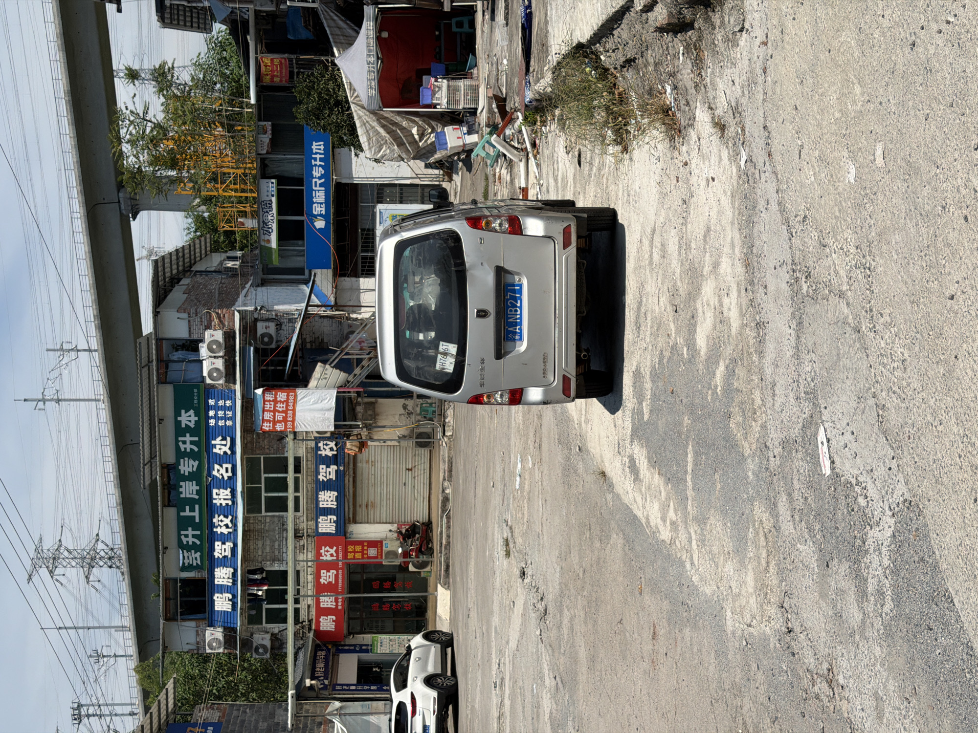 This image captures a street scene in the city of Jiangjin, China, under a bright, overcast sky. A silver minivan is parked in the foreground, facing away from the viewer, its license plate reading "A NB271". To the left of the minivan, a white SUV is parked.
The background features a row of buildings with multiple stories, adorned with various signs. One prominent green sign reads "茗升上岸专升本" which translates to "Ming Sheng Shang An Zhuan Sheng Ben" (likely referring to an educational institution or program). Below this, blue and red signs display Chinese characters, some of which appear to be advertisements for driving schools ("鹏腾驾校"). Air conditioning units are visible on the building facades.
A pedestrian overpass or bridge, supported by concrete pillars, spans across the upper portion of the image, with power lines running beneath it. To the right, a red tent structure with white trim is set up, with various items, including plastic crates and what appear to be stacked chairs, visible underneath and around it. The ground in front of the buildings is a rough, cracked asphalt surface, with some patches of weeds and debris scattered around. The overall impression is that of a slightly run-down urban environment, possibly a commercial or residential area with ongoing activities.
