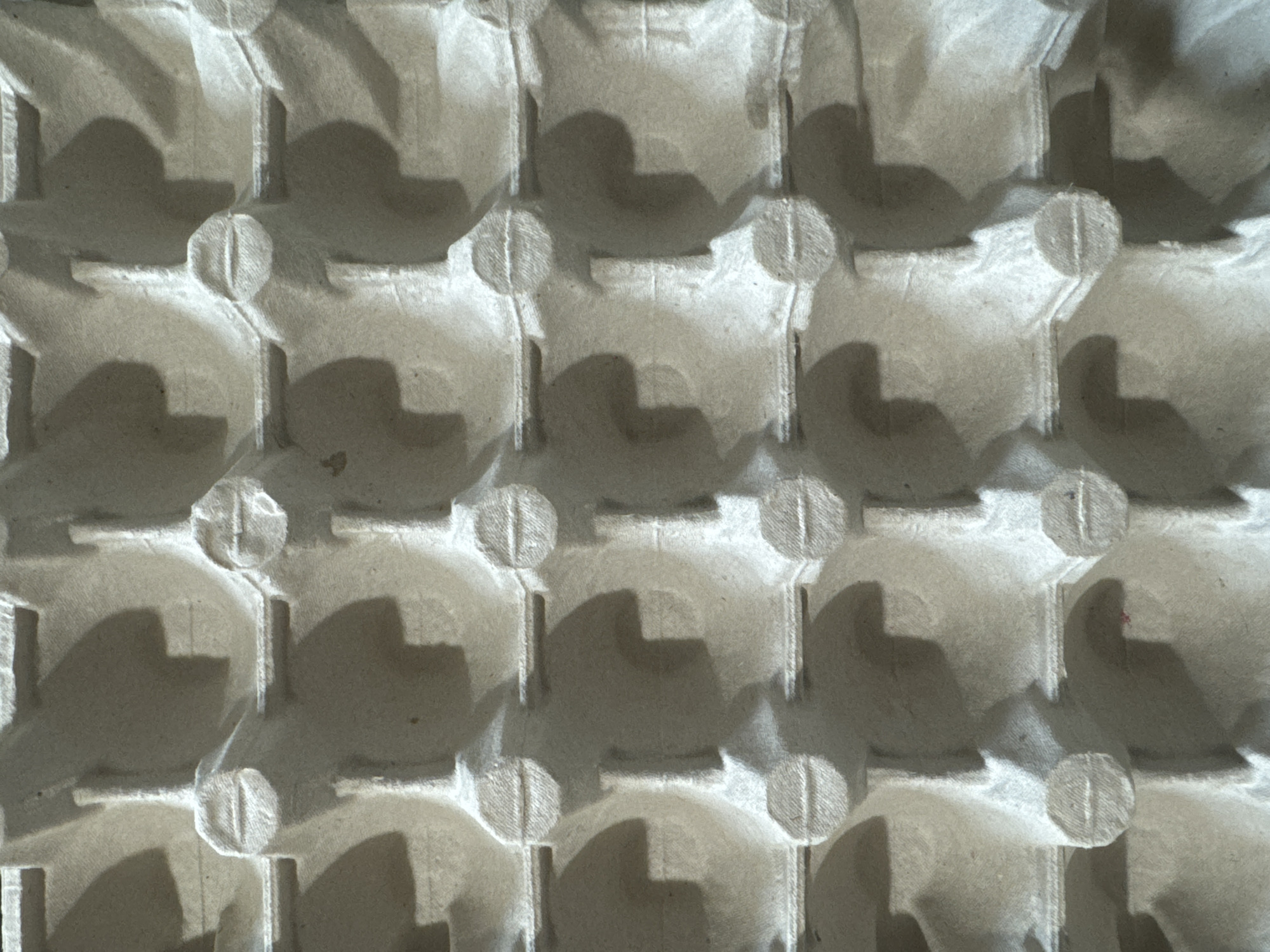 The image shows a close-up of a molded pulp fiber egg carton. The grid pattern consists of hemispherical depressions for holding eggs, connected by vertical and horizontal structural elements. Small circular pads are located at the intersections of the vertical and horizontal elements. Shadows are cast across the surface of the carton, creating varying shades of light and dark. The material is a uniform light gray-brown color. The exact location in the city of Chaikovsky, Russia is not determinable from the image.