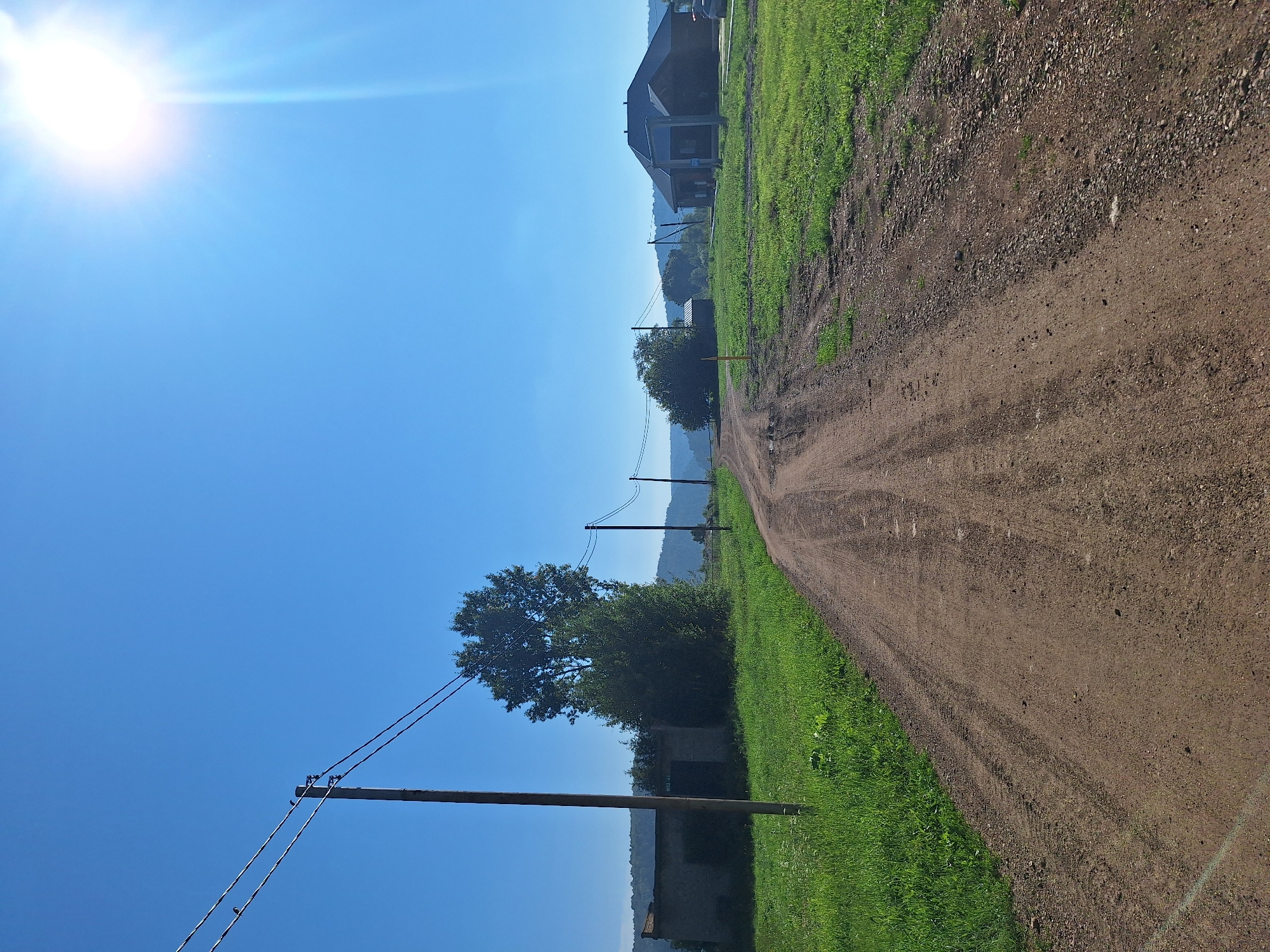 In Atamanovka, Russia, on a bright, clear day, a dirt road stretches into the distance under a vibrant blue sky. The sun shines brightly in the upper right corner, casting a warm glow. On the left side of the road, a long, low building with a dark facade sits amidst lush green grass, with a prominent wooden utility pole standing nearby. Further down the road, another utility pole is visible, followed by a cluster of trees and a dark, modern-looking house with a steep roof on the right side of the road. The road itself shows tire tracks, indicating recent vehicle passage. Rolling hills are visible in the background, softened by a light haze. The overall scene depicts a rural setting on a sunny day.