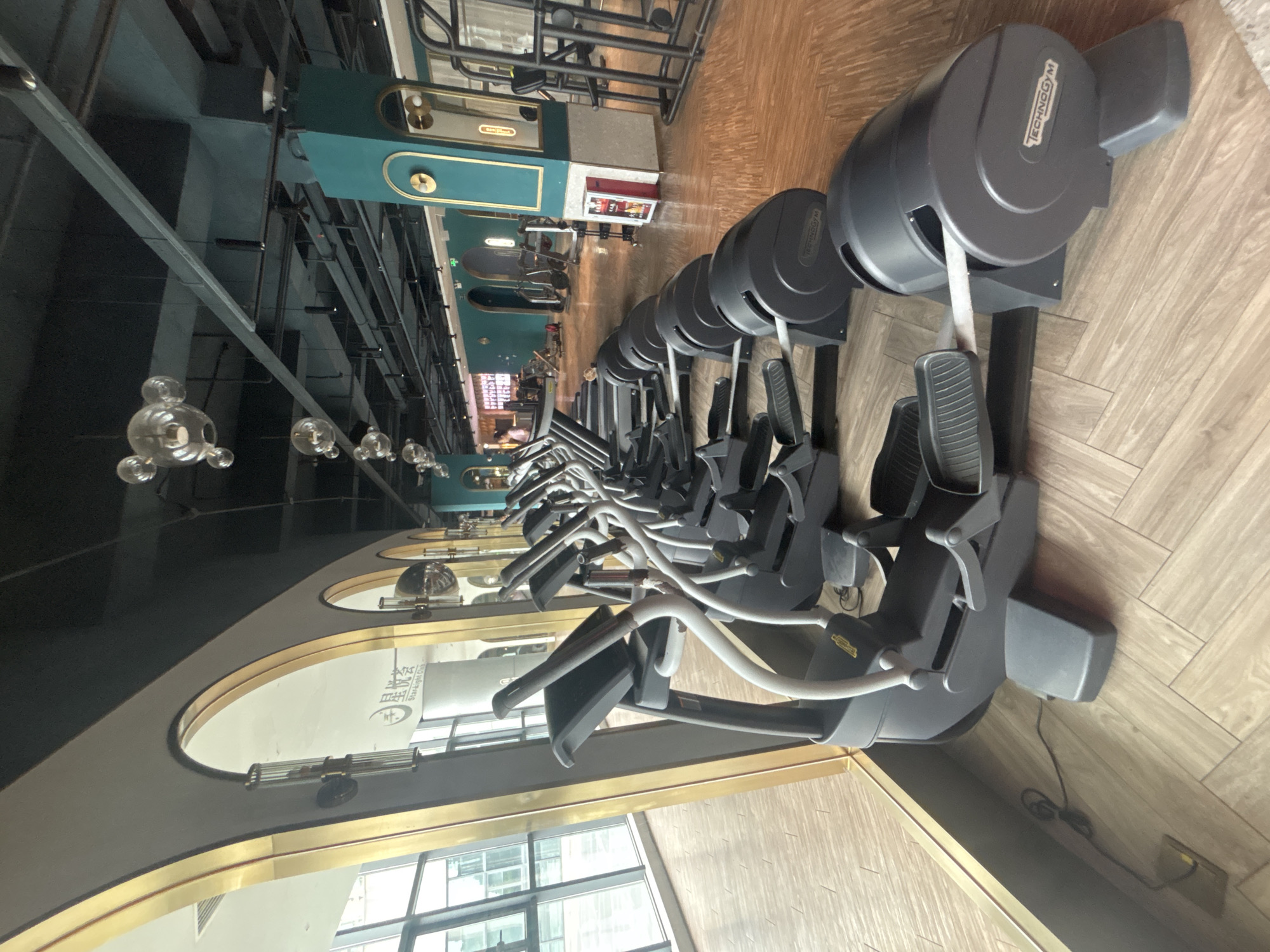 This image depicts the interior of a modern gym, likely located in Nanhai District, China, given the signage visible. The scene is dominated by a row of gray Technogym elliptical machines, positioned on a light wood-paneled floor. These machines are neatly arranged, facing towards the left side of the frame, with their pedals and handlebars visible. The gym's architecture features large arched windows with gold trim along the left wall, offering a glimpse of an exterior urban environment. The ceiling is dark and industrial, with exposed beams and track lighting, and decorative spherical light fixtures hang down. The background reveals more gym equipment and teal-colored accent walls with gold decorative elements, suggesting a sophisticated and well-maintained facility. No people are prominently featured in the foreground, but a few figures can be seen in the distance towards the right, engaged in their workouts. The overall impression is one of a clean, organized, and upscale fitness space.