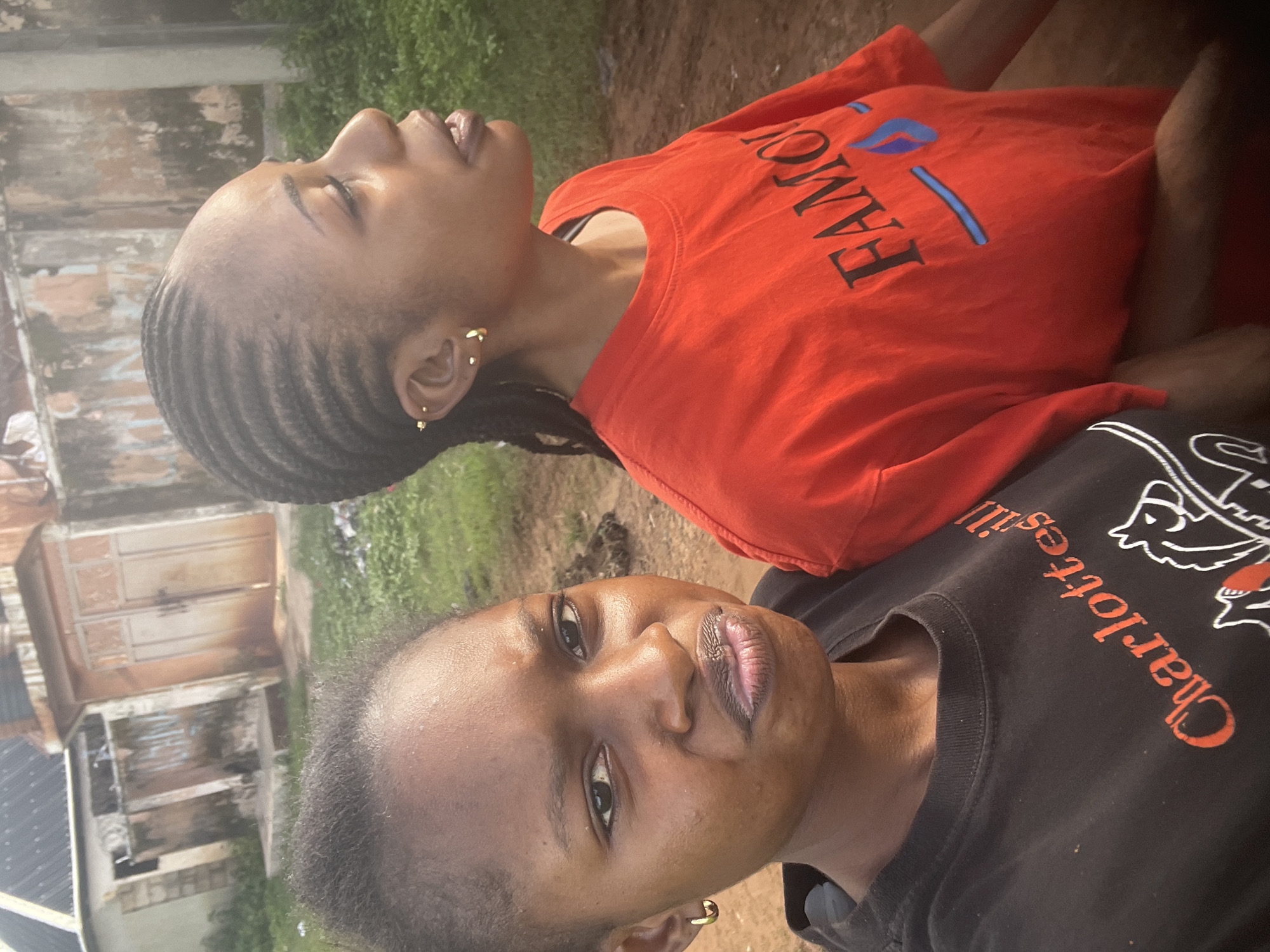Two young Black women are captured in a close-up, outdoor portrait. The woman on the left, with short hair and wearing a black t-shirt with "Charlottesville" and horse graphics printed in orange, looks directly at the camera with a slight frown. She has a gold hoop earring in her visible ear. The woman on the right, with braided hair, wears an orange t-shirt with the word "FAMOUS" printed in black. She is looking to the right of the frame. Her ears are adorned with small gold studs and a small hoop. In the background, a dirt path is visible with some sparse grass and a weathered, tan building with large, double doors and barbed wire fencing. The scene appears to be in Lafia, Nigeria, suggesting a location in a residential or semi-urban area. The lighting suggests it is daytime, possibly late afternoon due to the warm tones.