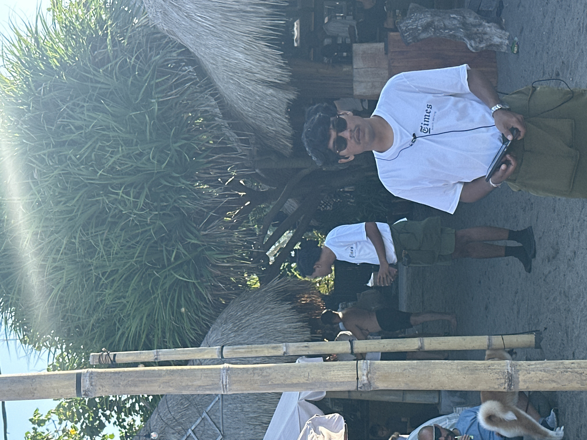 In North Kuta, Indonesia, two men stand near thatched-roof huts. The man in the foreground wears a white t-shirt with "Times" printed on it and sunglasses. He holds a phone in his left hand. Another man, also in a white t-shirt and shorts, stands behind him, holding a drink. A dog sits near a man in a blue shirt on the left side of the frame, partially obscured by bamboo poles. The environment appears to be a beach or outdoor cafe setting with sandy ground.