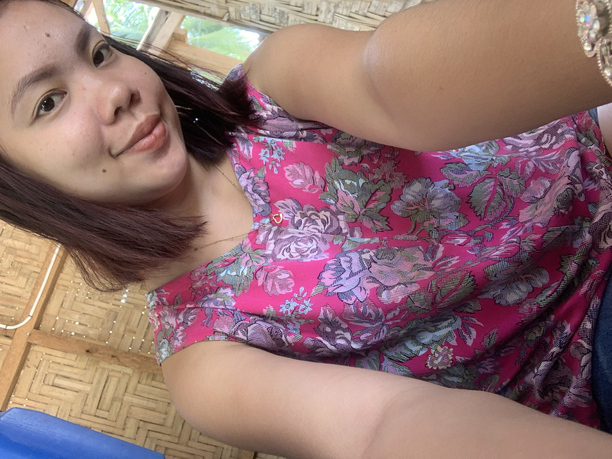 A young woman with dark hair and a light complexion takes a selfie in a room with walls made of woven bamboo. She is wearing a pink floral shirt and a delicate gold necklace with a heart-shaped pendant. Her left arm is extended toward the camera, obscuring part of her body and the background. The lighting is natural, casting soft shadows. The background also shows glimpses of what appears to be vegetation through a window. The overall setting suggests a rural or informal dwelling. There are no obvious signs of violence or distress. The image does not provide any specific information that could pinpoint its location within Kiblawan, Philippines, beyond the general description of the dwelling.