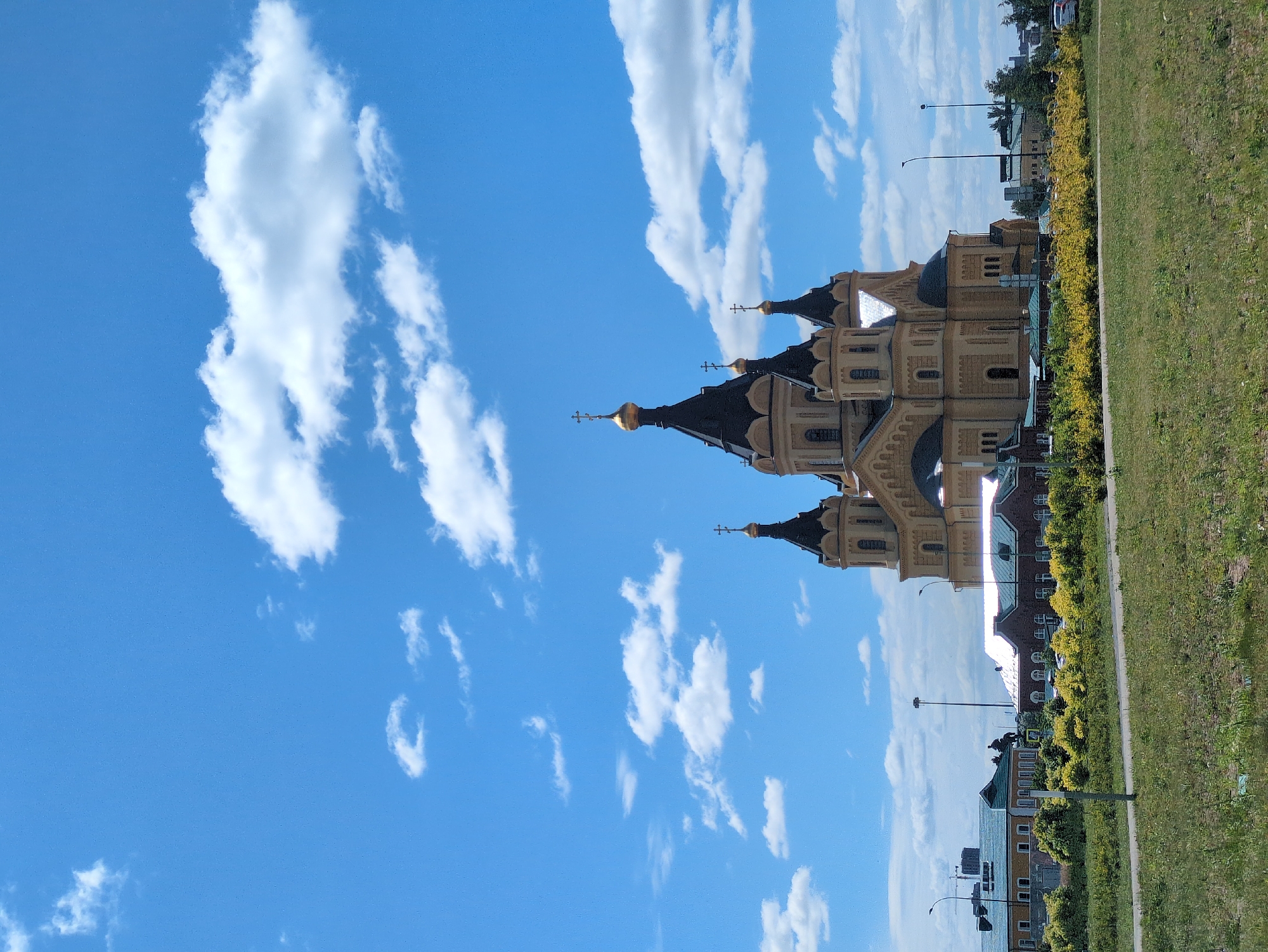 The image depicts the Alexander Nevsky Cathedral in Nizhny Novgorod, Russia, on a clear day with a bright blue sky dotted with fluffy white clouds. The grand cathedral, with its distinctive golden domes and intricate architecture, stands prominently in the background. In the foreground, a well-maintained grassy area leads to a pathway, with a line of vibrant yellow bushes separating it from a street with some buildings and lampposts. The overall scene conveys a sense of peacefulness and architectural grandeur.