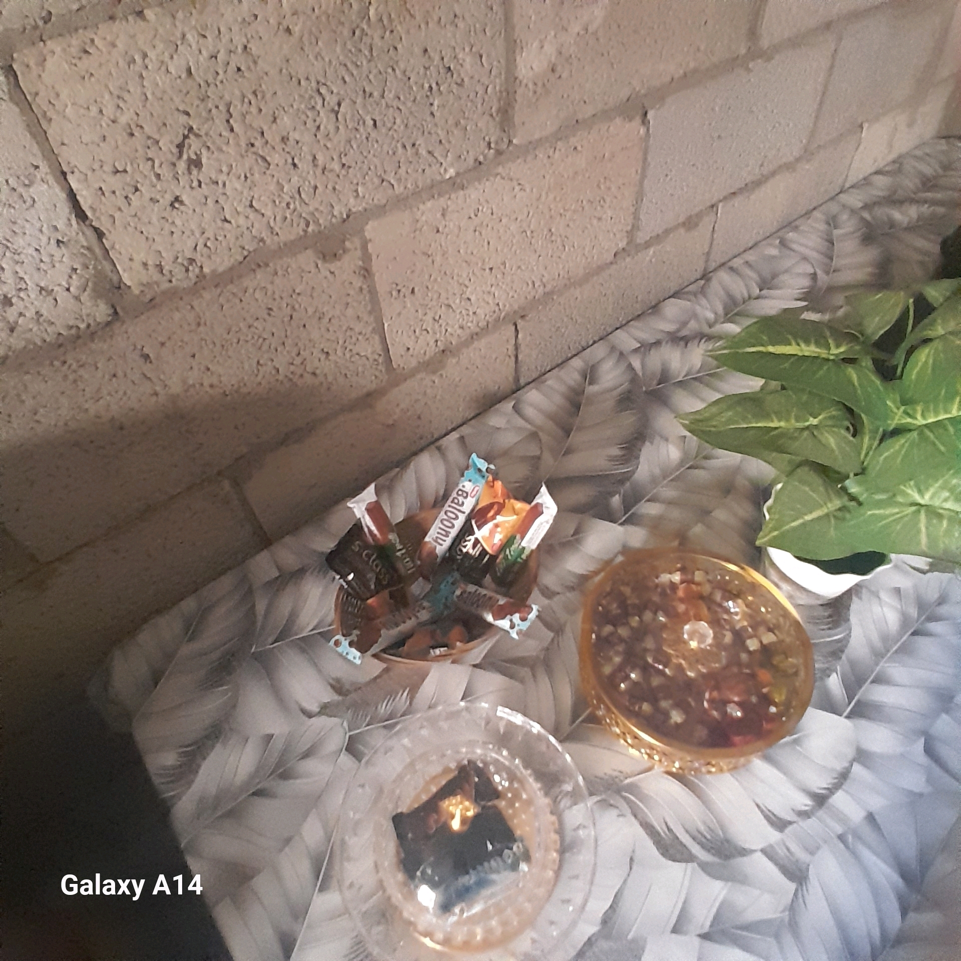The image displays a close-up, high-angle shot of a table covered with a grey and white patterned tablecloth. On the table, there are several objects, including a small pile of candy wrappers, a decorative gold bowl filled with small objects, a clear glass dish with what appears to be a dark object inside, and a potted plant with green leaves. The background consists of a textured, light-colored brick wall. The text "Galaxy A14" is visible in the lower left corner of the image. There are no people or visible actions occurring in the scene. The environment appears to be indoors, possibly a room within a house or a shop.
