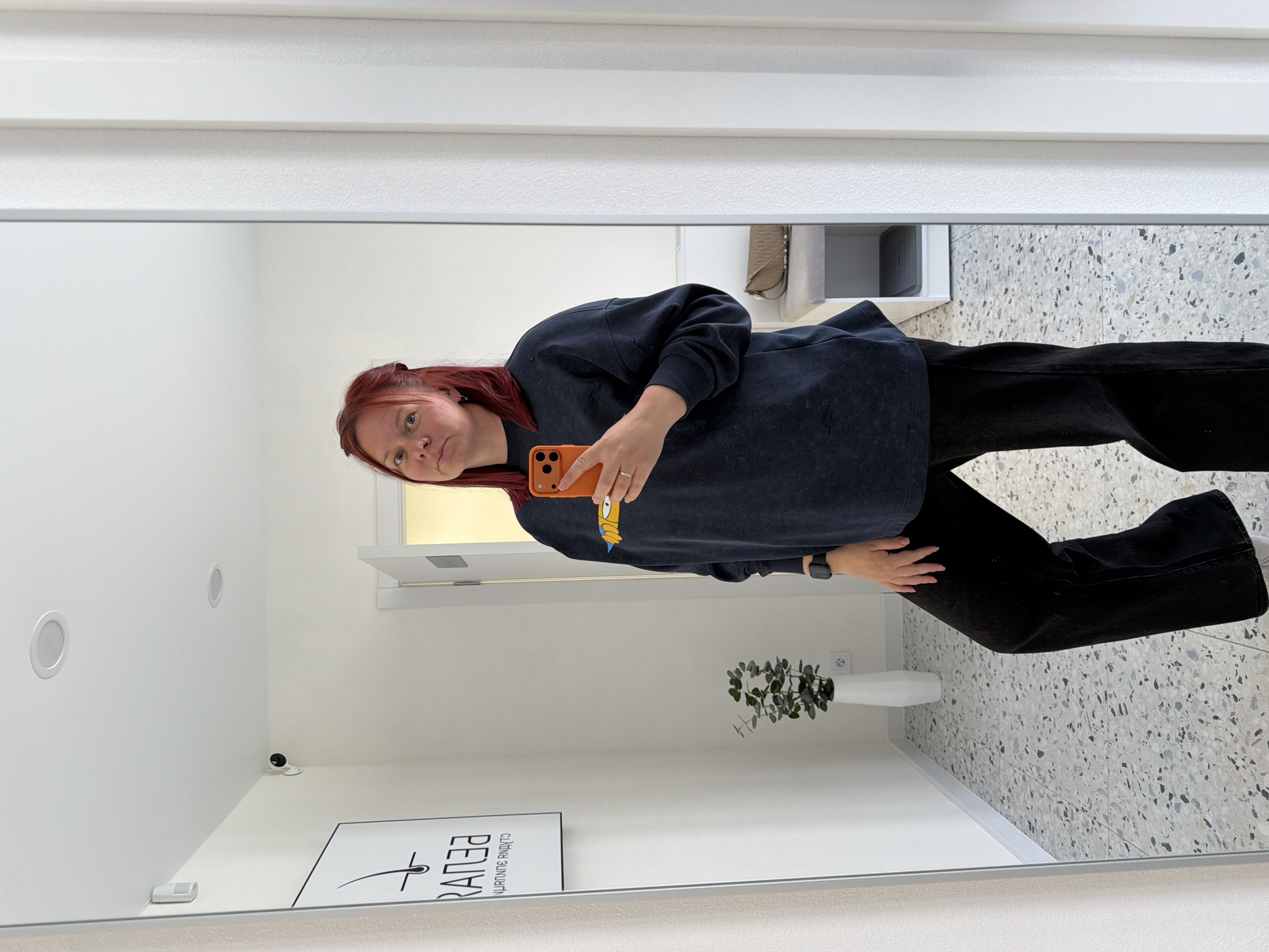 A reddish-haired person stands in front of a mirror, holding a smartphone with an orange case. The person is wearing a dark sweatshirt and black pants. The reflection shows a clean, brightly lit interior space. The floor has a speckled pattern. There is a vase with greenery, a framed advertisement or sign, a security camera, and other interior elements visible in the reflection. Based on the sign in the reflection, the location may be in the city of Chaikovsky, Russia.