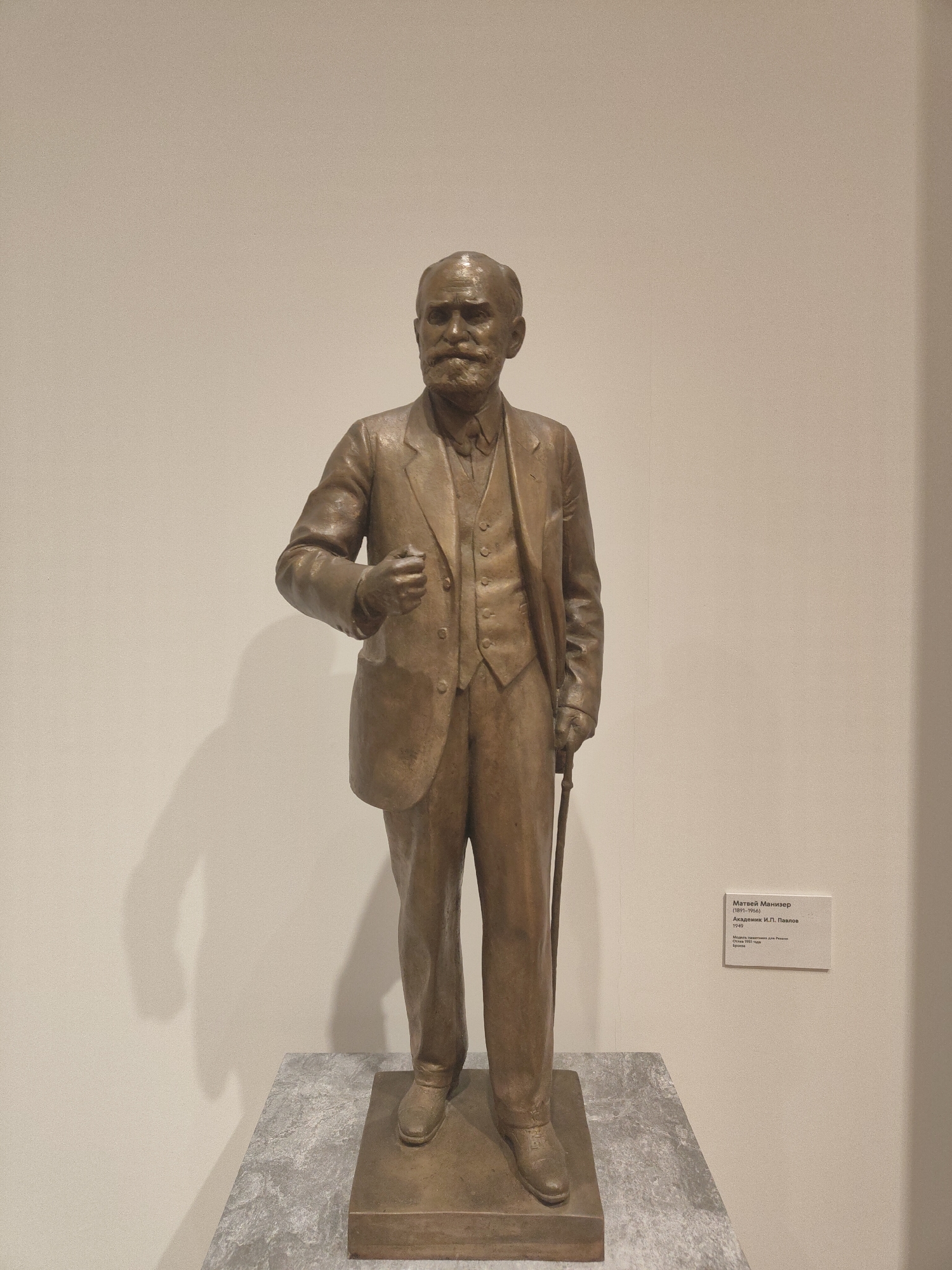A full shot captures a bronze statue of a man in a suit, cane in hand, standing on a gray marble pedestal. The statue is situated indoors, possibly in a museum, against a plain, light-colored wall. To the right of the statue, there is a white information plaque detailing the artist and subject matter in Russian. The statue depicts a man with a receding hairline and a prominent mustache and beard, dressed in a three-piece suit. His left hand is slightly clenched, while his right-hand rests on a cane. The statue is located in an unknown location in the city of Unknown, Unknown.