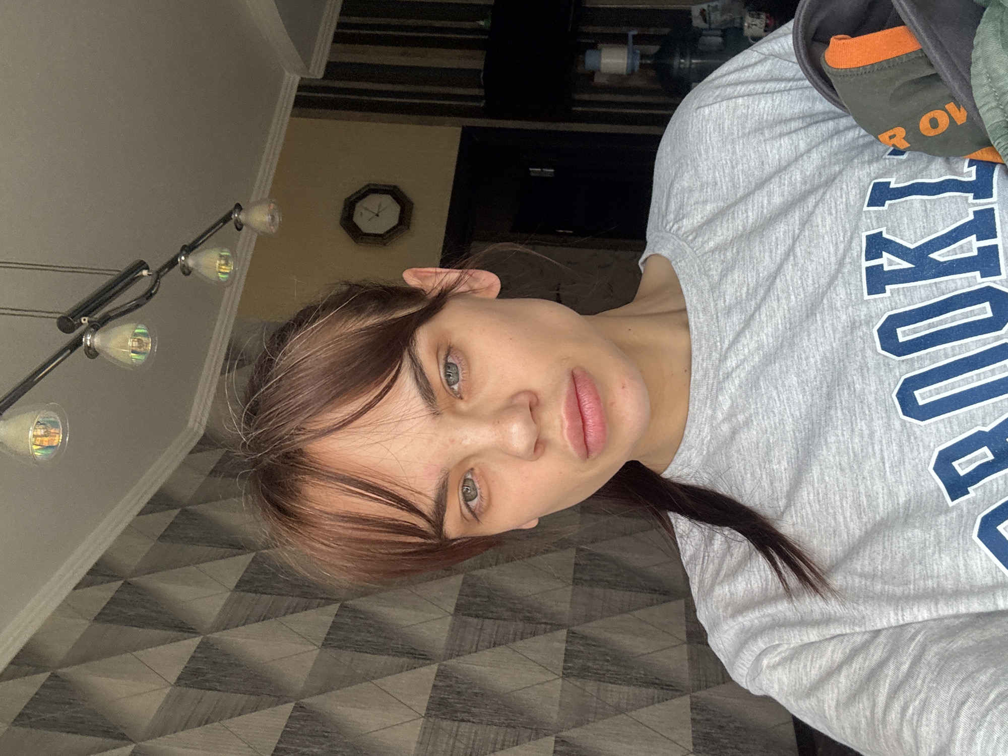 A woman is facing the camera. She has brown hair with auburn streaks in her bangs. She is wearing a gray long-sleeved shirt with navy blue lettering. A portion of an orange collar and olive green fabric is visible over her right shoulder. A chandelier with five lights hangs from the ceiling. A clock is visible on the wall behind her. The floor has a geometric gray-and-white pattern. The location is Rostov-on-Don, Russia.