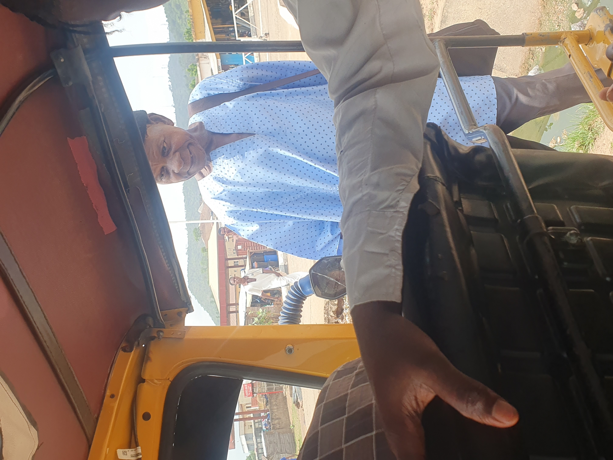 The image, taken from inside a yellow auto rickshaw (keke), shows an elderly man in a light blue patterned shirt riding as a passenger. The photo appears to have been taken while the keke was in motion, possibly within the city of Lokoja, Nigeria, as can be inferred by the presence of buildings and the terrain visible through the keke's open sides. Another person is visible partially, whose arm and hand are in the foreground. In the background, other buildings and people are visible outside the auto rickshaw.