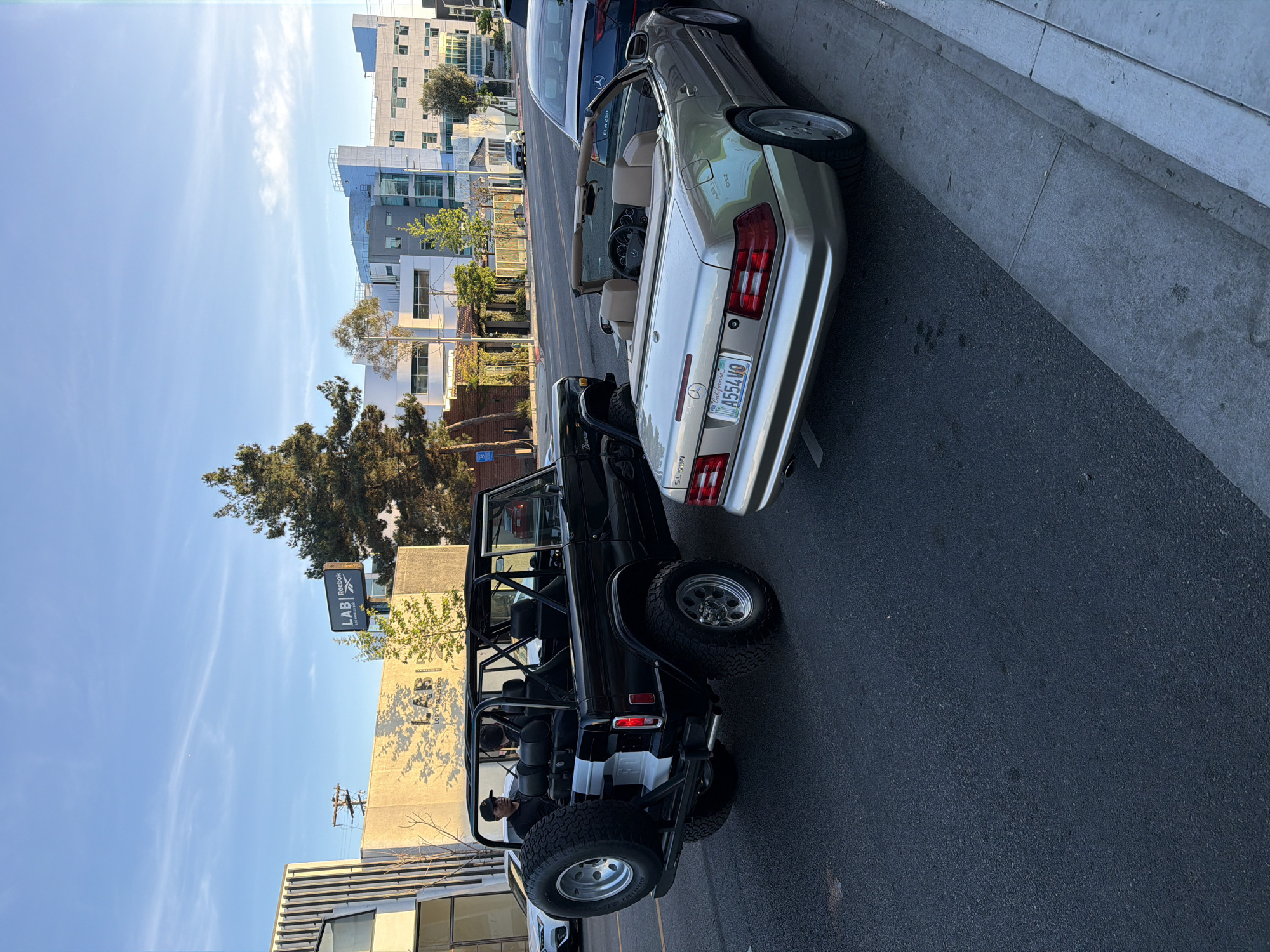 The image captures a street scene in West Hollywood, United States of America, featuring parked vehicles. A black Bronco is parked on the left, its rear facing the viewer. Beside it, a silver Mercedes-Benz convertible is parked with its rear partially visible. The buildings in the background consist of a mix of modern and older architectural styles. The sky is blue with few clouds. Signage for "LABI" is visible in the background. There are no discernible people in the scene aside from the Bronco occupant and possibly the Mercedes occupant, although the details are unclear. The environment suggests a sunny day in a typical urban setting.