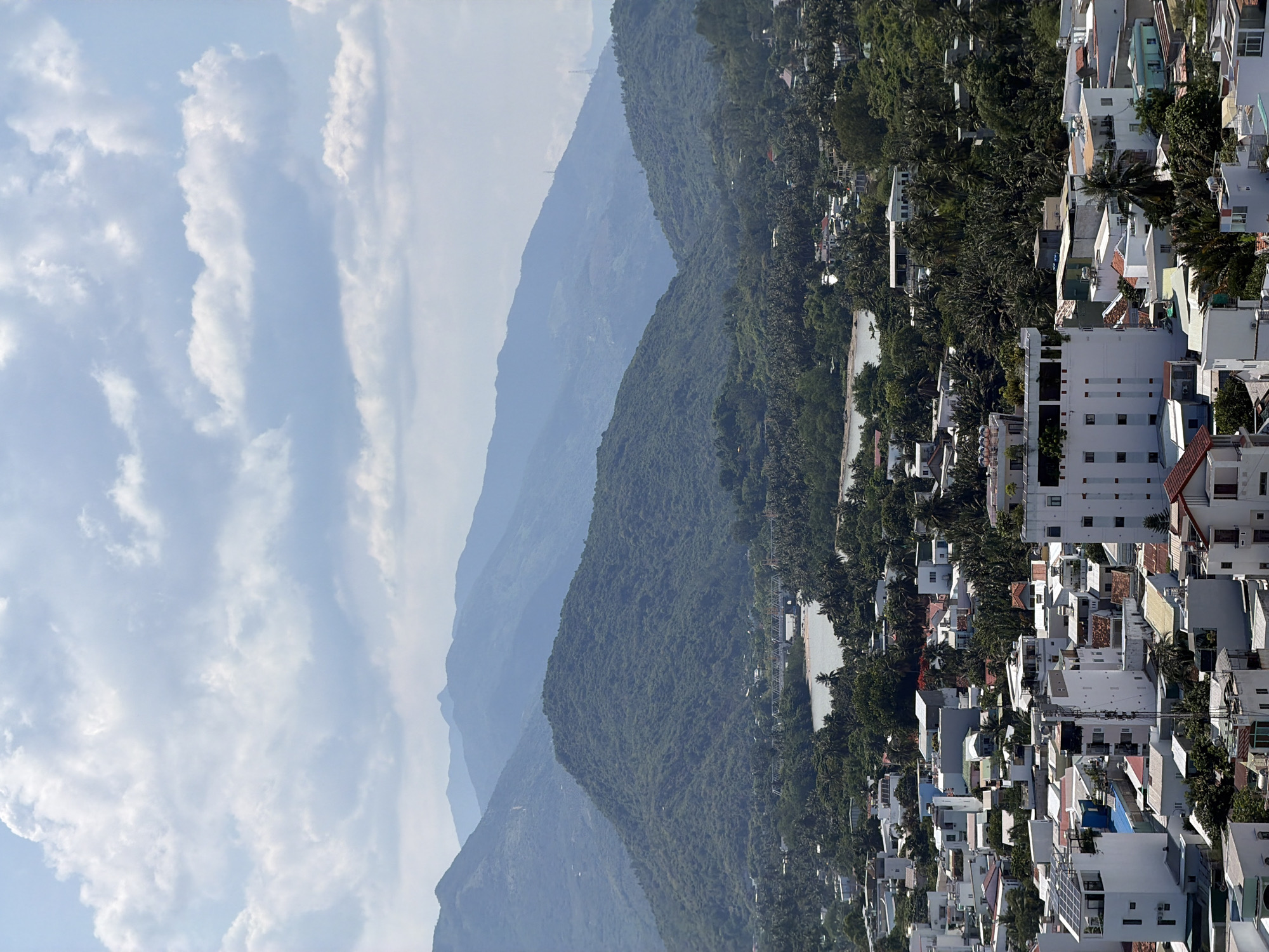 This is a vertical, eye-level, outdoor photograph taken on a bright, partly cloudy day overlooking a coastal city in Nha Trang Ward, Vietnam. The foreground is dominated by a dense urban landscape of white, multi-story buildings with flat roofs, interspersed with lush green palm trees and other vegetation. Solar panels are visible on the roofs of some buildings. In the mid-ground, a river or body of water flows through the city, crossed by a white metal bridge with an arching structure. Beyond the river, the landscape transitions to densely forested, green hills, which are the foothills of several layers of progressively distant, hazy blue mountains that fill the background and fade into the sky. The sky is a pale blue, with scattered white, fluffy clouds. No people are visible in the image. The scene captures a view of urban development nestled between natural mountainous terrain and a waterway, indicative of a coastal city in Vietnam.