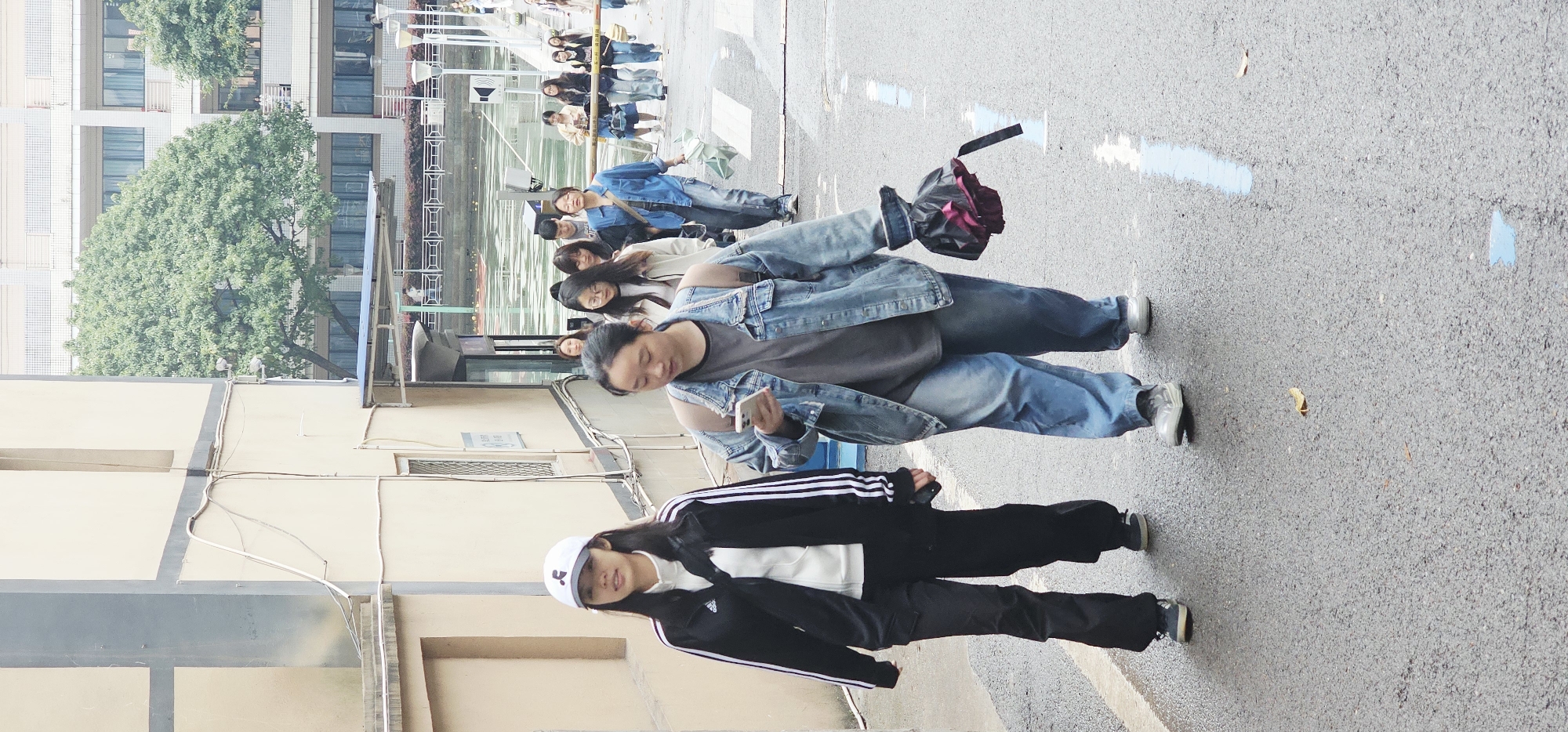 A group of people are standing on a gray asphalt surface next to a building in what appears to be an urban setting. The location may be Yuzhong District, China. The building is light colored with multiple windows and appears to be of moderate height. In the foreground, two individuals are prominently visible. One is wearing a black track suit with white stripes and a white baseball cap. The other is wearing a gray shirt, jeans, and a denim jacket. She is holding a smartphone. Other people can be seen standing in the background, near a body of water and a safety railing. Blue markings are on the asphalt surface.