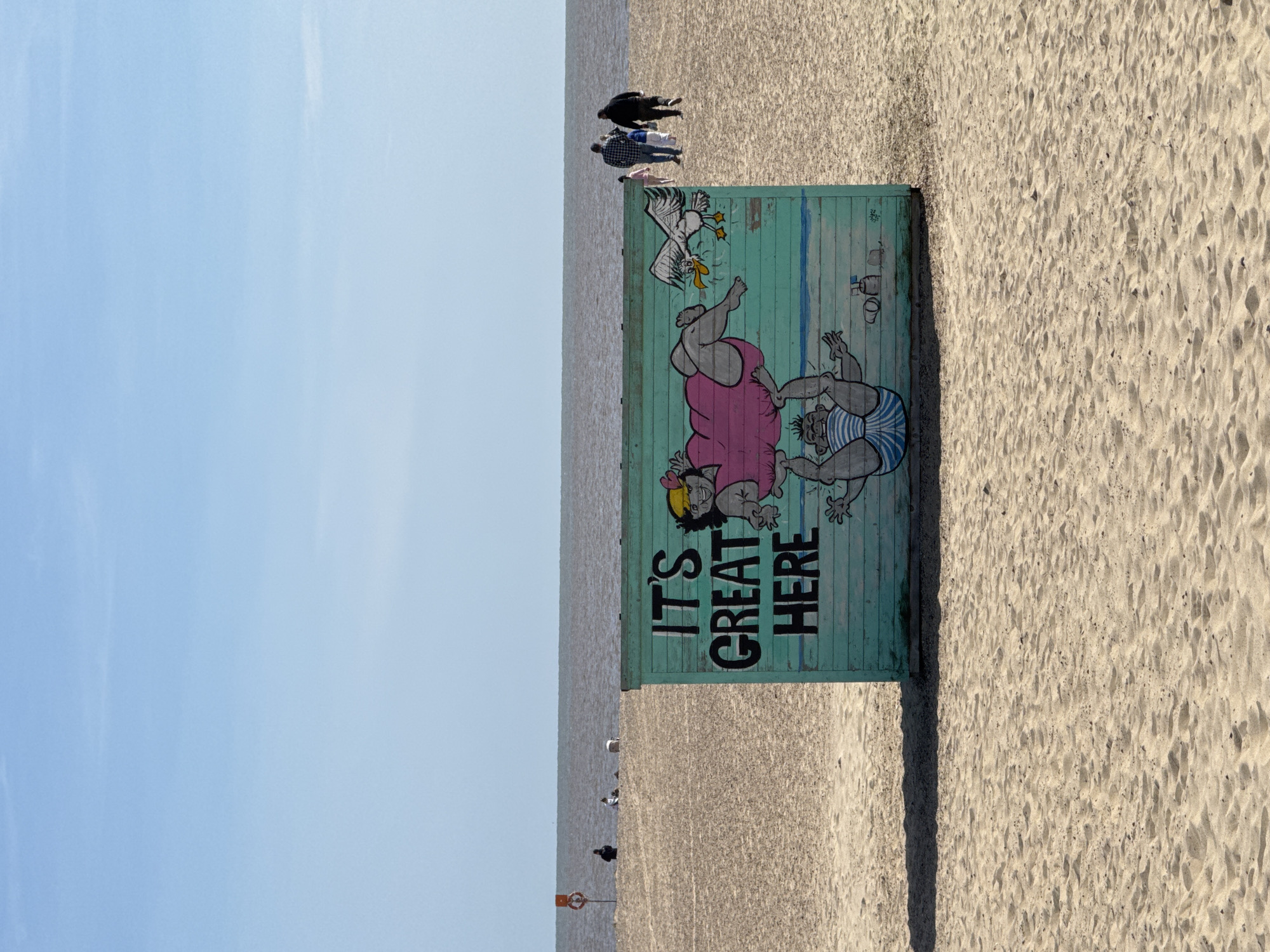 The photograph, taken at Great Yarmouth, United Kingdom, presents a seaside scene. The horizon line is located in the centre of the shot, with a clear sky in the upper left. A stretch of light brown beach occupies the lower right section.
In the foreground, a wooden beach hut stands prominently. The hut has an illustration painted on it depicting cartoon figures of people and a bird, accompanied by the text "IT'S GREAT HERE".
In the background, several people are visible on the beach near the horizon. The sea is a grey colour, with a few objects visible in the distance, possibly buoys.