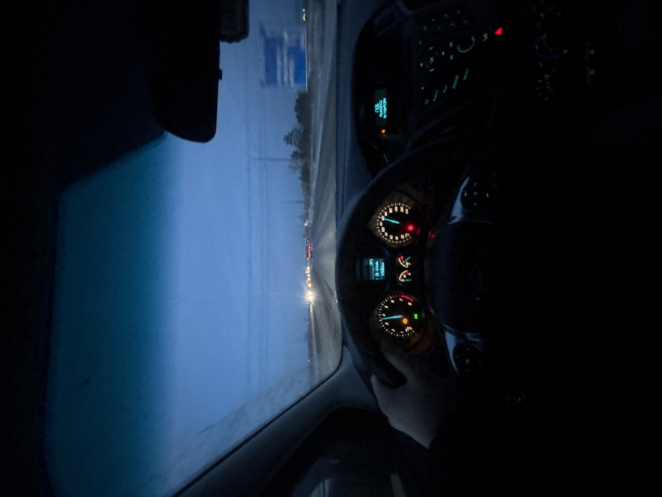 The image displays a first-person perspective from inside a moving vehicle on a road in Terent'yevo, Russia, during low-light conditions, indicative of dusk or dawn. A human left hand is positioned on the black steering wheel at approximately the 9 o'clock position. The vehicle's instrument cluster is illuminated: the speedometer indicates a speed between 100 and 110 km/h, with an active blue indicator light. The tachometer shows a green indicator light and a blue engine coolant temperature light. A central digital display within the cluster registers an ambient temperature of 3°C and a distance reading of 17336.0 km. The central console's infotainment system is partially visible to the right, illuminated with blue and green controls. The rearview mirror is visible at the top center. The windshield shows frost or condensation on its upper left portion and appears generally obscured by precipitation or residue. Outside the vehicle, a multi-lane road, appearing damp or lightly covered in snow/slush, stretches into the distance. Other vehicles are visible ahead, identifiable by their illuminated headlights and taillights. A large blue road sign with white, illegible text is present on the right side of the road. Utility poles or power lines line the roadside. The sky overhead is a uniform dark blue, suggesting an overcast environment.