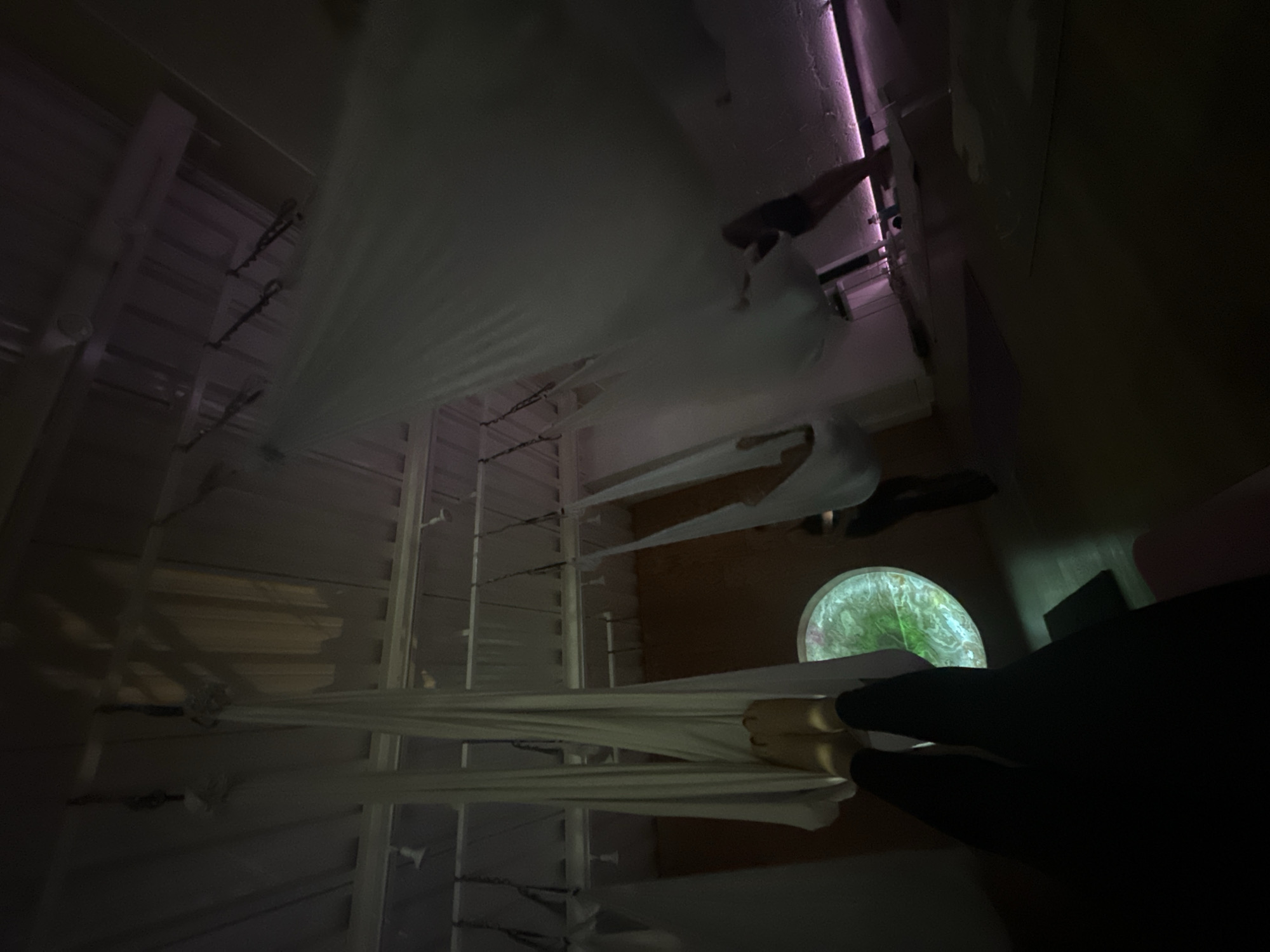The image depicts a room interior with an unconventional perspective, likely from an aerial or suspended position. The focus is on a series of horizontal white fabric strips stretched across the room, possibly part of an art installation. The background includes elements of a window with a circular frame, through which greenery is visible. The person wearing black tights is partially visible in the photo. The lighting is dim, with areas of shadow and some sources of illumination, including what appears to be a fluorescent or LED light strip. There are white vertical window blinds. The location of the scene is not discernible from the photograph. The image does not provide any information to identify the location of Puebla, Mexico.