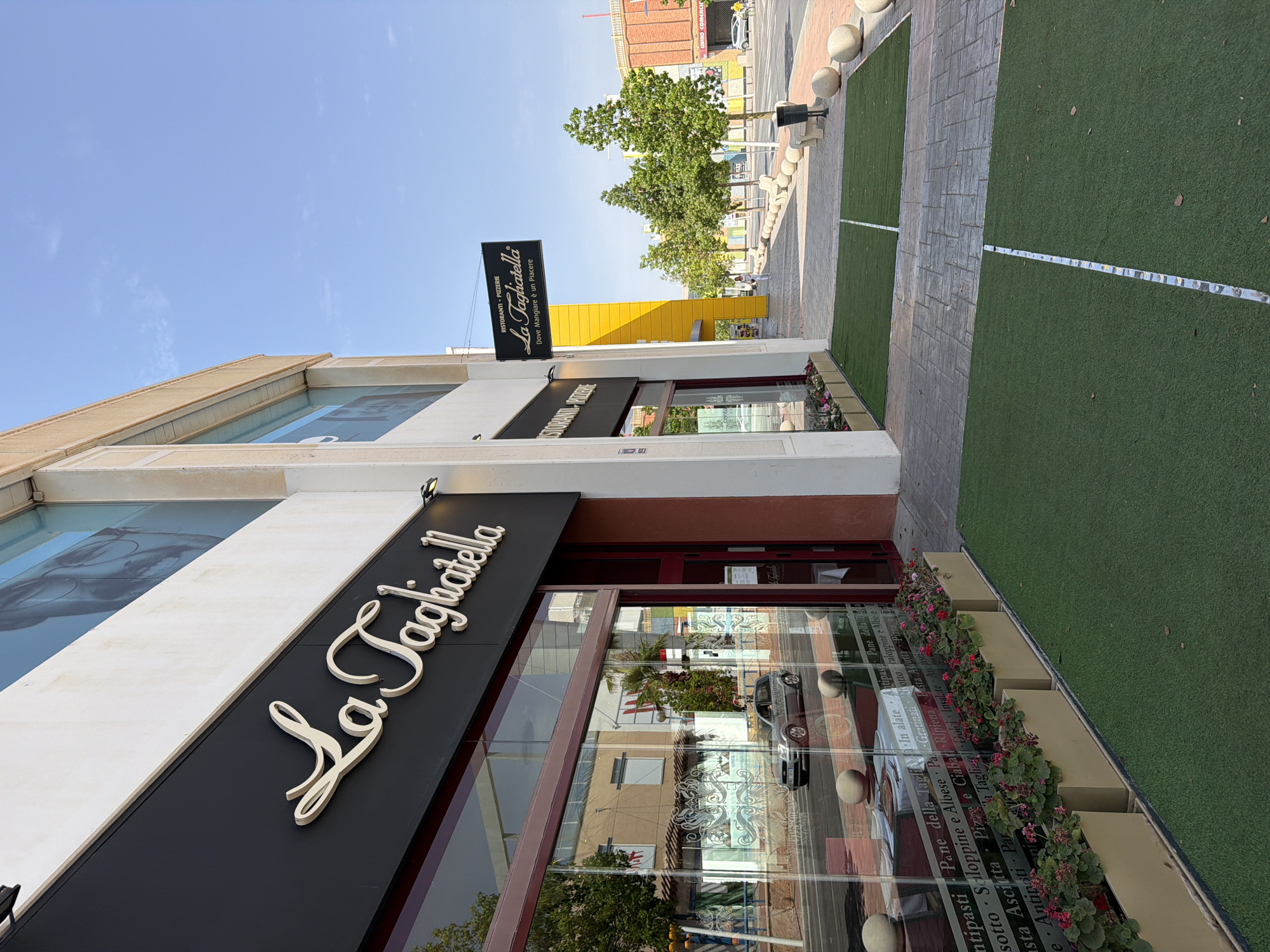 The image shows the exterior of a "La Tagliatella" restaurant in La Cala, Spain. The restaurant has a large glass storefront with the name "La Tagliatella" in prominent, stylized lettering above the entrance. A secondary sign hanging from the building reads "Ristoranti • Pizzerie La Tagliatella Dove Mangiare è un Piacere".
In front of the restaurant, there is a sidewalk with a patterned stone paving and several large, round concrete bollards. Sections of artificial turf are laid out along the sidewalk, marked with white lines. A few trees with green foliage are planted along the sidewalk, and there are planters with pink and green flowers in front of the storefront.
Through the glass storefront, tables are visible inside the restaurant, set with white tablecloths and place settings. A silver car is reflected in the glass, driving on the street in front of the restaurant. The street itself is paved, and there are other buildings and trees visible across the street. The sky above is a clear blue with a few wispy clouds. A person is walking in the distance on the sidewalk.