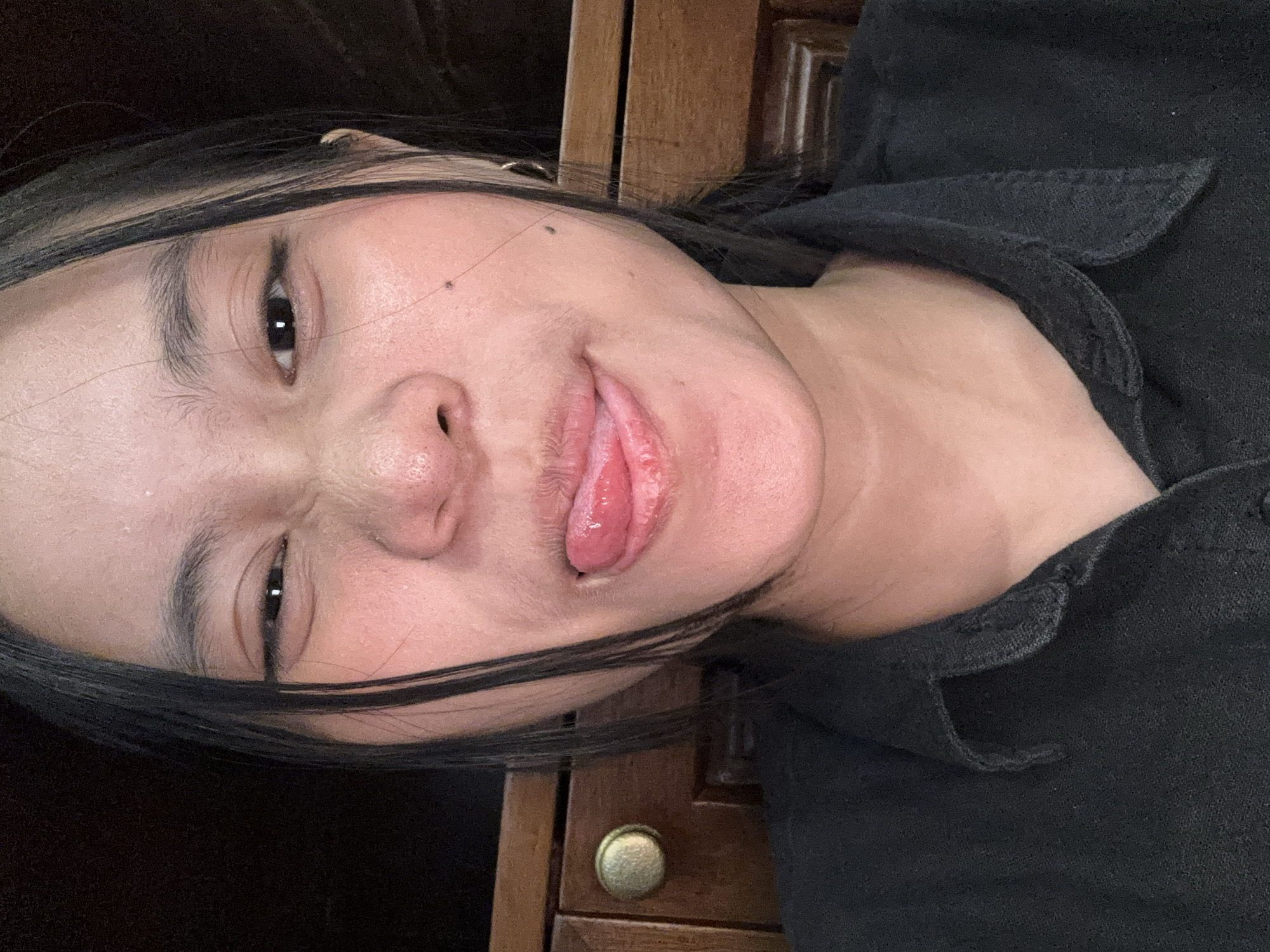 The image is a close-up, frontal selfie of a young woman with dark hair and light-colored streaks, possibly highlights, framing her face. She is wearing a dark, collared shirt. Her eyes are partially closed, and her tongue is sticking out, with a glossy appearance, suggesting she might be moistening her lips or playing for the camera. Her skin appears flushed, and there are a few visible pores and a mole on her right cheek. The background is dimly lit and appears to be an indoor setting, with the textured wood of a cabinet visible to the right. No specific location in South Jakarta, Indonesia is discernible from the visual cues in the image.