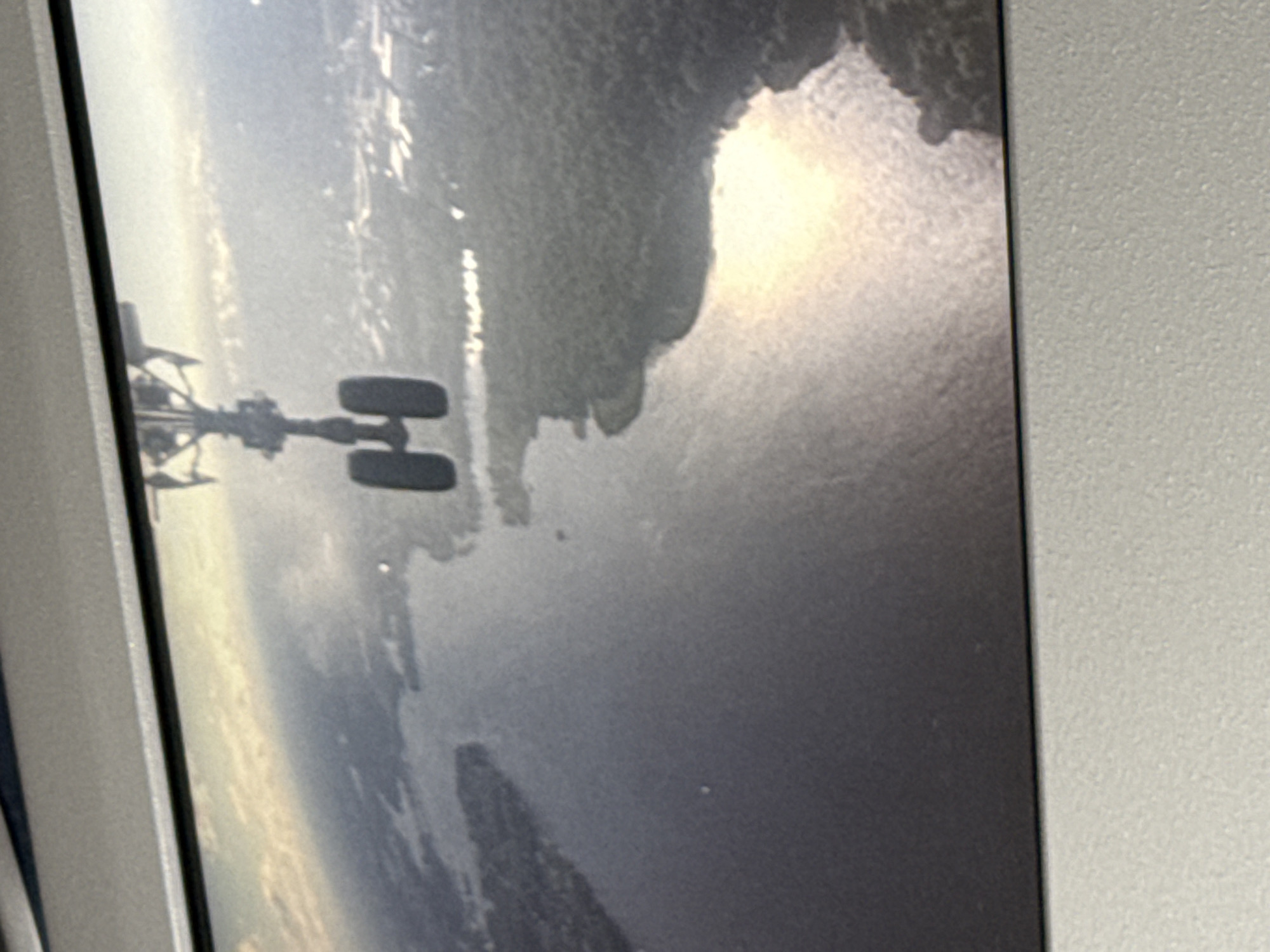 The image displays a perspective from an aircraft, looking down at a body of water and a treed landscape. The aircraft's landing gear, consisting of two wheels, is prominently visible in the foreground, suggesting the plane is either preparing for landing or has just taken off. The water surface reflects the sunlight, creating a bright, shimmering effect. The surrounding land is covered in dense forestation, with some indications of urban development or infrastructure in the distance, possibly a town or city. The scene is consistent with an aerial view over a river or lake system adjacent to a populated area. While the image itself does not contain explicit markers of the city of Rakovo, Russia, such a view could be encountered when approaching or departing from an airport near a body of water like the Lake of Rakovo, if such a geographical feature exists.