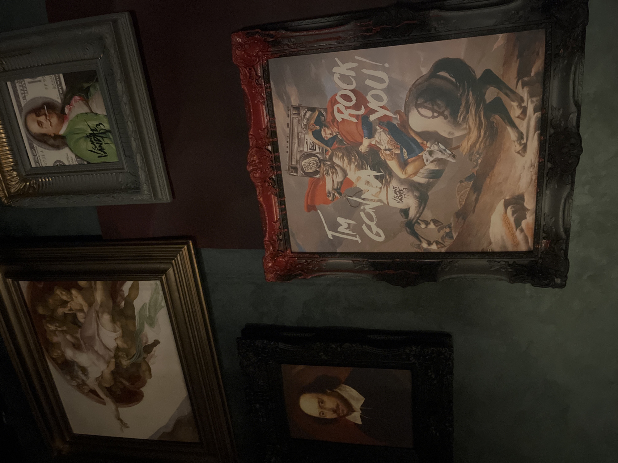 The image depicts a wall adorned with several framed artworks. In the upper left corner, a framed painting reminiscent of Michelangelo's "The Creation of Adam" is visible. To its right, a framed print features a stylized portrait of Benjamin Franklin superimposed on a one-hundred-dollar bill, with graffiti-like text below. Below the Benjamin Franklin print and to the left of the center, a framed portrait of William Shakespeare is displayed. Dominating the lower right quadrant is a large, framed artwork with a red, ornate frame. This central piece is a modern interpretation of Jacques-Louis David's "Napoleon Crossing the Alps," with the figure of Napoleon riding a horse, a boombox on the horse's back, and stylized text that reads "I'M GONNA ROCK YOU!" superimposed on the image. The overall impression is of an eclectic collection of art displayed in a dimly lit interior space, possibly a bar, restaurant, or private residence. The location in Stavropol, Russia is not discernible from the visual elements present in the image.