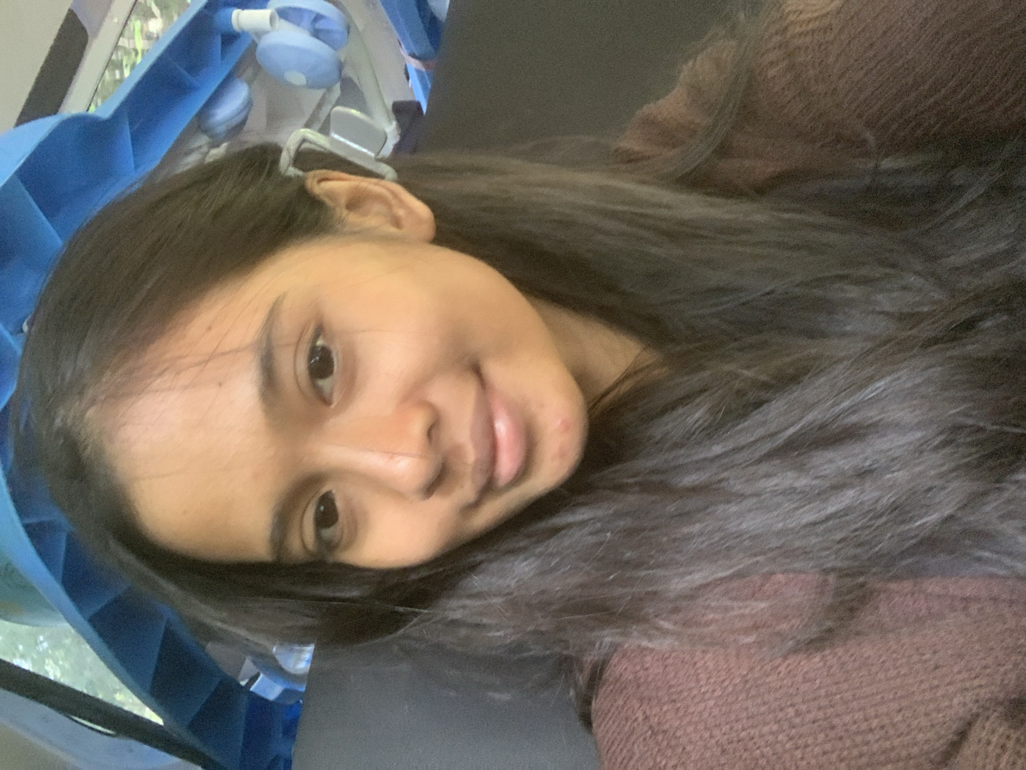 A close-up selfie shows a young woman with dark hair. She is wearing a brown sweater. Behind her, there's blue medical equipment. The location is not discernible.