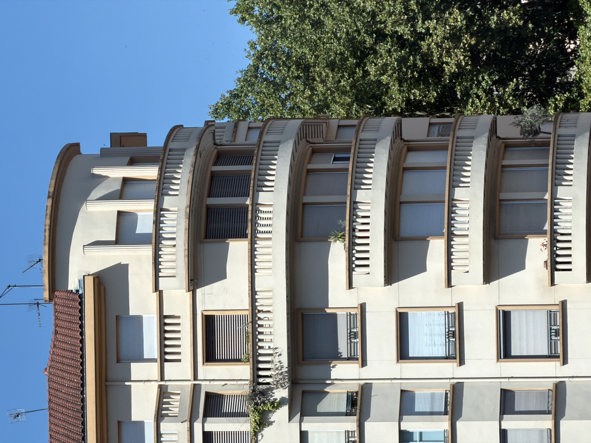 The image shows a section of a multi-story residential building in Lyon, France. The building has a distinctive rounded corner with balconies on each level. The balconies are enclosed with decorative railings. The building's facade is light-colored, possibly stucco or concrete, with darker accents around the windows and balconies. The windows are fitted with roller blinds. A red tiled roof is visible at the top of the building, along with television antennas. A large, mature green tree is positioned to the right of the building, partially obscuring some of its facade. The sky is clear and blue, indicating a sunny day. There are no people visible in the image. The architectural style appears to be from the early to mid-20th century. The building is situated in a developed urban area, as suggested by the presence of multiple floors and the contrasting natural element of the tree.