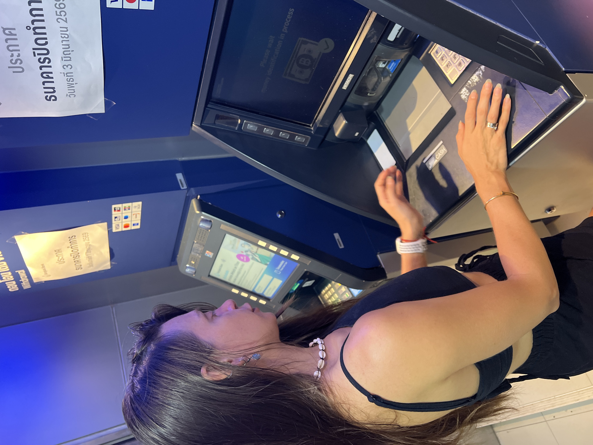 The photograph shows a person using an ATM. The person is a woman with long, dark hair wearing a black top and a seashell necklace. She is inserting a card into the ATM and has her other hand on the keypad. The ATM is a Hitachi model. The ATM is in a public space, and there is a sign in Thai next to it stating that the bank is closed on Wednesday, June 3, 2569. There is also another sign next to that sign. The location is likely in Baan Bo Phut, Thailand. The ATM screen displays the message "Please wait, money identification in process" and a picture of a bank note.