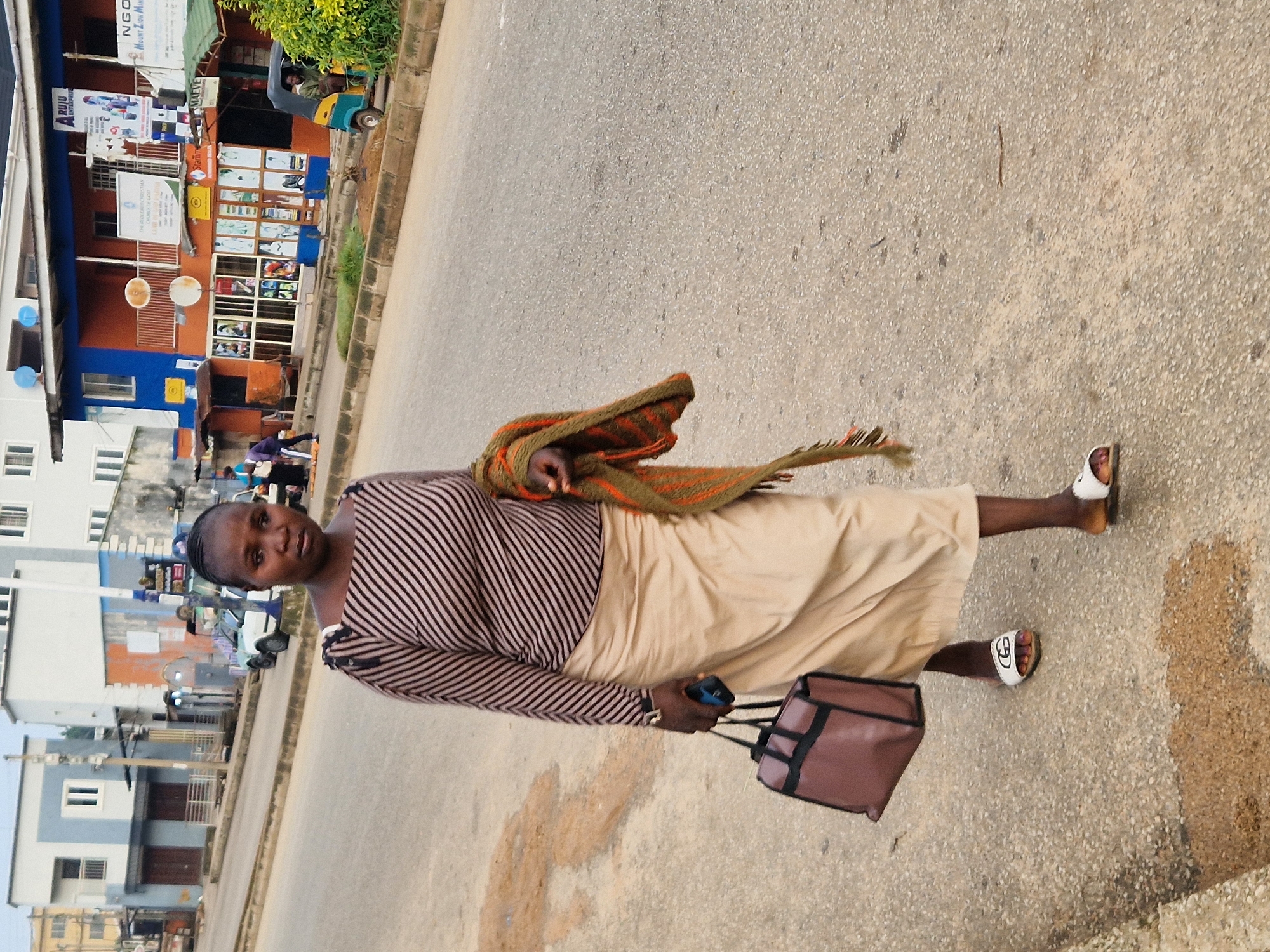 A full-shot, eye-level, outdoor, daytime, street-level, vertical-format, medium-detail photograph shows a woman walking on a paved street in Kabong, Nigeria. The woman, who is the primary subject, is of Black African descent and has dark skin, dark braided hair, and is wearing a striped long-sleeved top and a beige skirt. She is carrying a brown bag in her left hand and a draped, colorful shawl or scarf over her right arm. She is wearing white slides on her feet. In the background, there are several buildings, a white car, a tricycle, and other people. The buildings appear to be commercial establishments and residential structures. The street is paved with asphalt and shows some signs of wear and tear, with a few puddles of water. The overall lighting suggests an overcast or early morning/late afternoon setting.