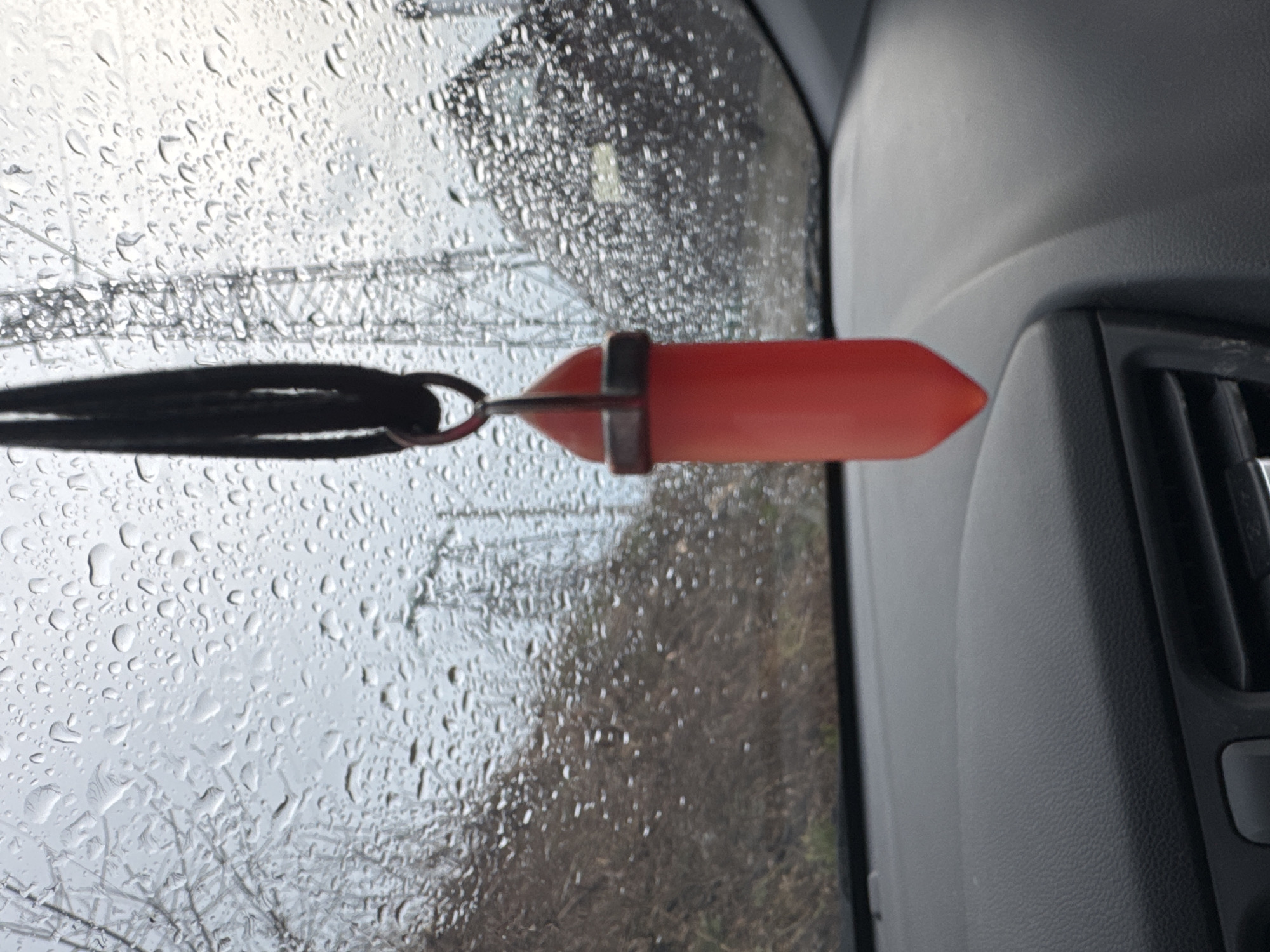 The image shows a red pendant hanging from a black cord, suspended in front of a car window covered in raindrops. Outside the window, blurred trees and possibly power lines are visible. The interior of the car is visible on the right side of the image, revealing a dark gray or black dashboard and part of the car's ventilation system. The location is likely Chaikovsky, Russia due to inference from the prompt. The scene appears to be taken from inside a car on a rainy day.