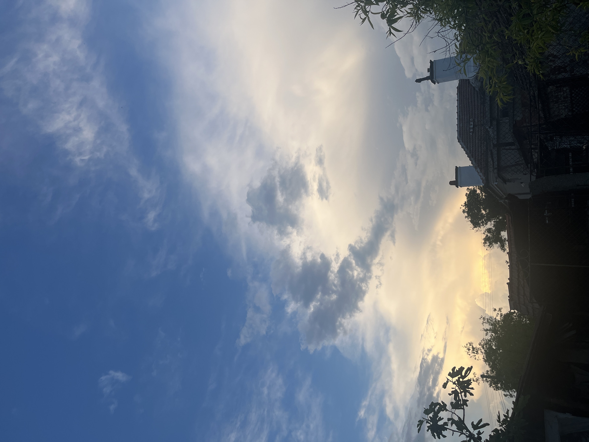 The image captures a view upwards, likely in Lozarevo, Bulgaria, showing a sky with a mix of blue and cloud formations. To the right, the roof of a building is visible, including two chimneys, partially obscured by trees and foliage. A fence is also visible adjacent to the building. The sky transitions from a clear blue to cloudy areas with shades of gray and white, with the light suggesting either sunrise or sunset.