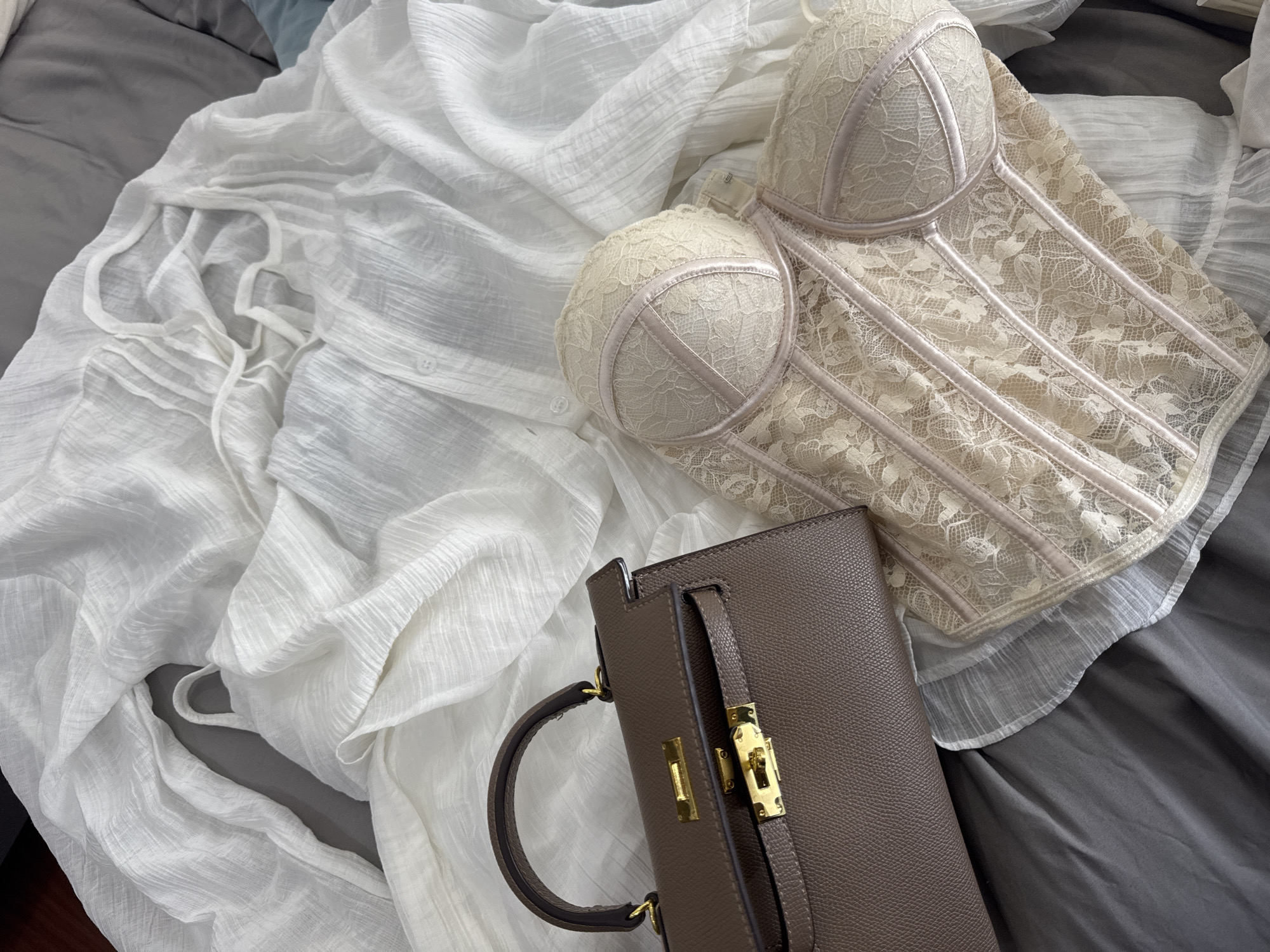 The image shows a close-up of various items on a surface. Dominating the composition are a beige lace bustier, a gray leather handbag with gold hardware, and a white button-down shirt. The objects are arranged on a bed with gray sheets. The location in the city of Đà Nẵng, Vietnam is not directly apparent.