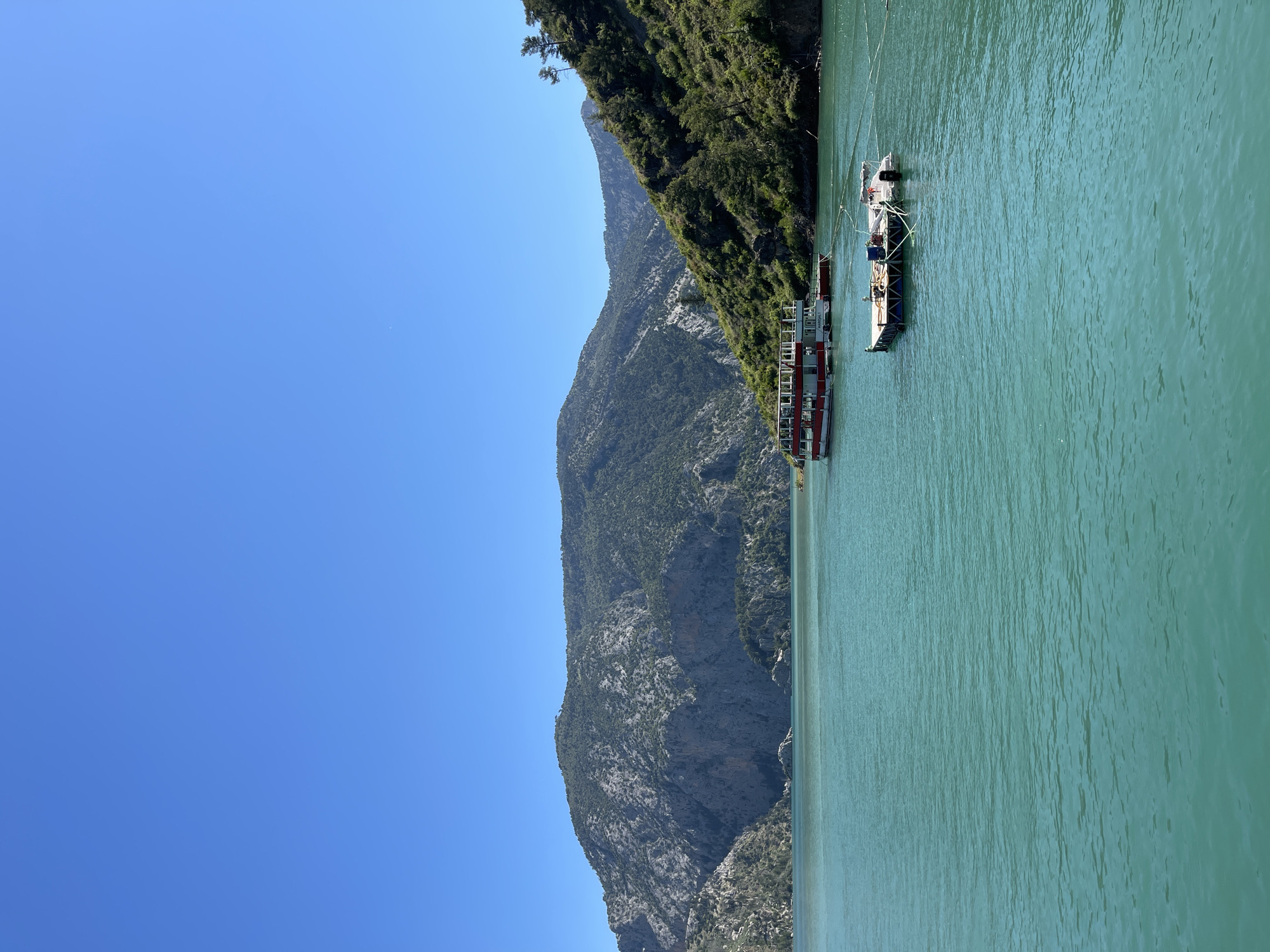 A vertical, medium shot shows a body of water with mountains in the background and two docked boats, taken during daylight hours. The sky above is clear blue. The mountains are rocky with some green vegetation. The water is a muted green hue and has some ripples and texture. The two boats are tied together. The boat closest to the foreground is a red and white tour boat with multiple levels. The second boat is a smaller, pontoon-style boat with metal framing and some equipment. The background includes the location in the city of Unknown, Unknown.