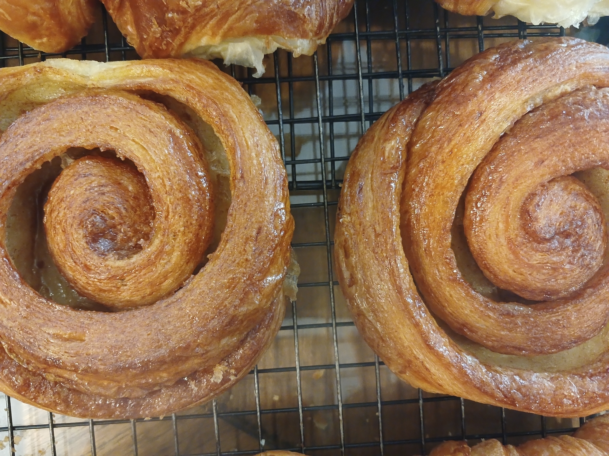 The image shows two spiral-shaped pastries, possibly cinnamon rolls or similar viennoiseries, sitting on a wire rack. Their golden-brown surface appears glossy, suggesting a glaze or butter wash. Some croissant-like pastries are also visible in the background. The scene suggests a bakery or kitchen setting. There is no indication that this scene is specifically located in Buenos Aires, Argentina.