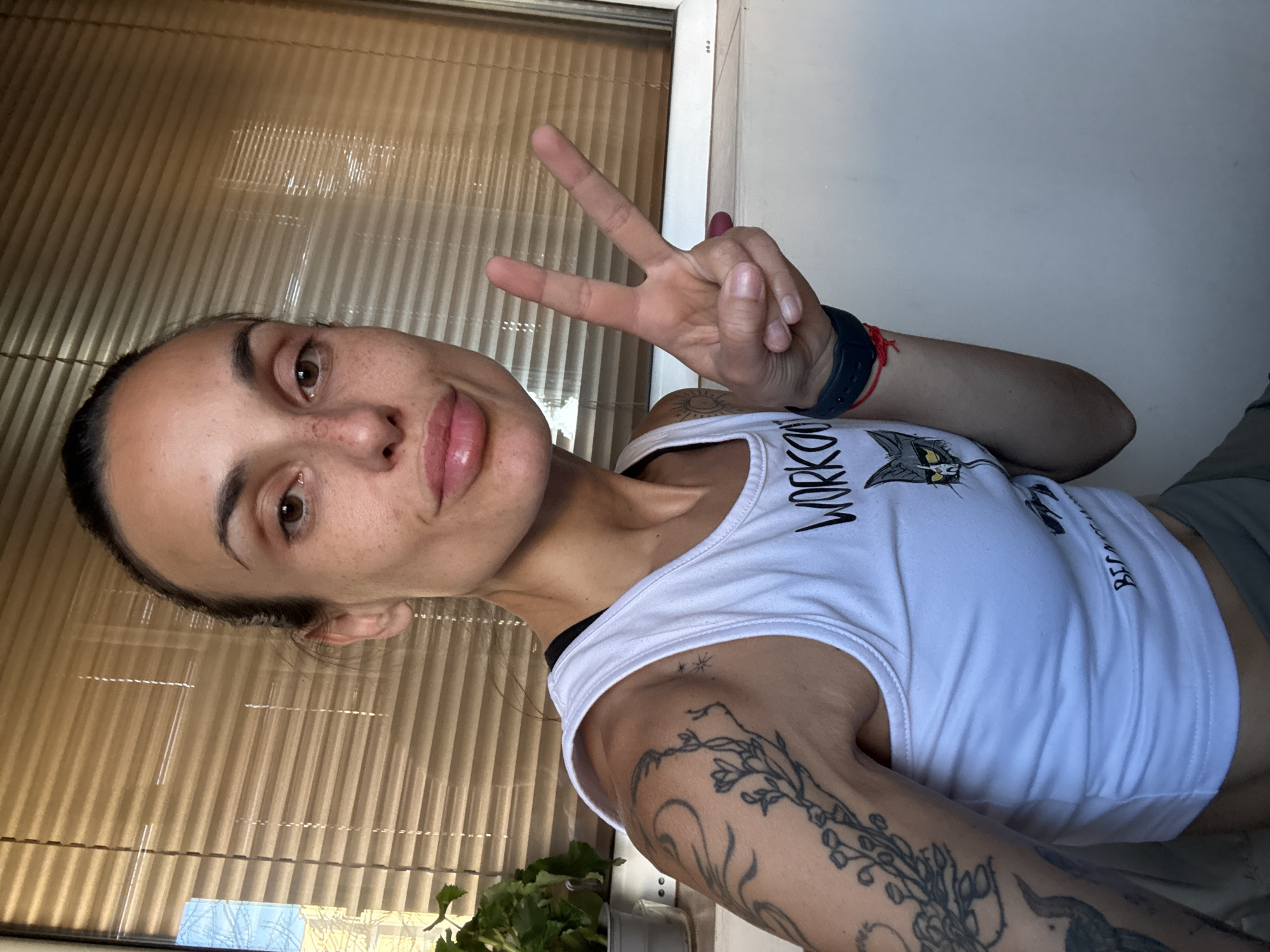 A young woman with dark hair pulled back and prominent freckles is taking a selfie in front of a window with horizontal blinds. She is wearing a white tank top with a graphic print of a cat and the word "WORKOU". On her right arm, she is making a peace sign with two fingers extended. She has several tattoos on her left arm. A green plant in a gray pot is visible to the left of the window. The setting appears to be indoors, possibly a residential building, with a plain white wall to the right of the woman. There is no specific indication of the location being in Pleven, Bulgaria.