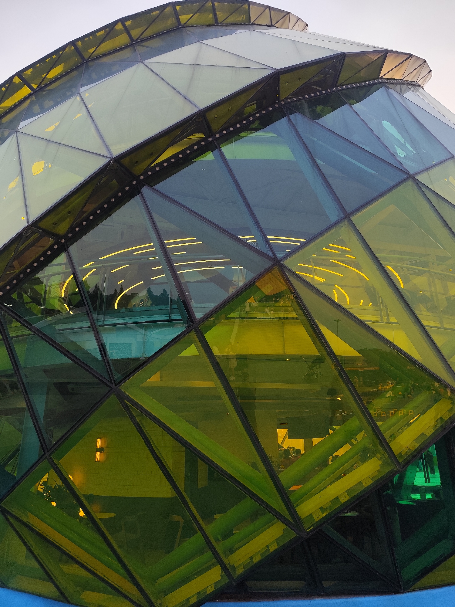 The image displays a close-up, upward-angled view of a modern, multi-faceted glass dome structure located at the Dalat Flower Garden in Da Lat, Vietnam. The building's exterior is constructed from numerous triangular and trapezoidal glass panels, predominantly tinted in shades of yellow-green, dark green, and lighter clear/bluish hues, interconnected by a dark structural framework. A horizontal band of small, round, white lights is visible along a perimeter structural element between two sections of the dome. The background sky is uniformly light grey. Through the varied glass panels, the interior of the structure is visible, illuminated by several curved, bright yellow linear lights arranged on the ceilings, casting reflections and patterns. Distant reflections of trees or foliage are also apparent on the glass surfaces. Inside, indistinct furniture, including tables and chairs or stools, can be discerned, along with a baby stroller visible through a lower pane. A blue-colored base or decorative element is visible at the bottom edge of the structure. No identifiable individuals or specific actions are clearly visible.
