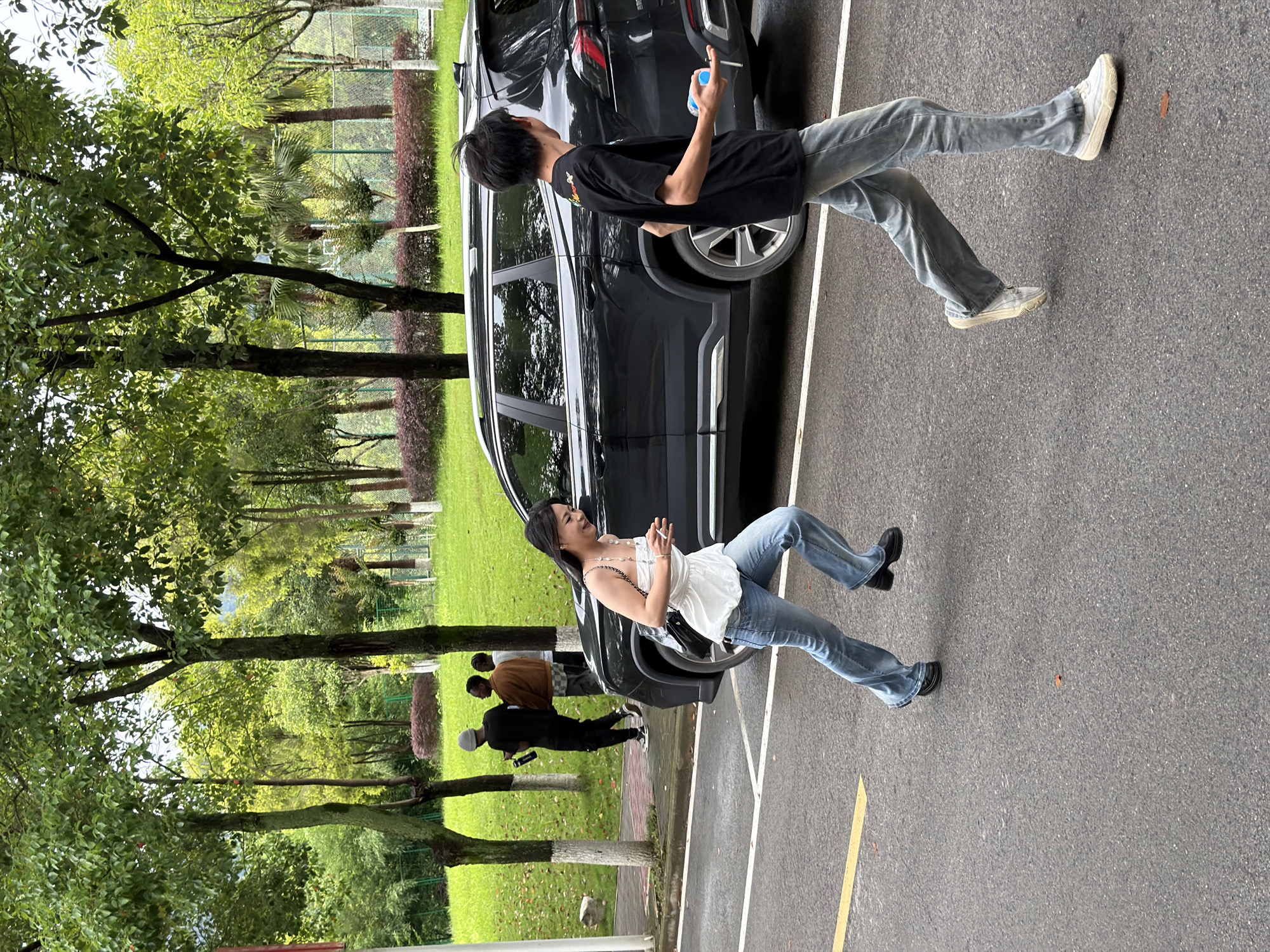 In Jiangjin, China, on a paved road bordering a grassy park with trees, a woman in a white top and blue jeans runs past a dark SUV. To the right of the SUV, a young man in a black t-shirt and light blue jeans walks away from the camera, holding a blue bottle. In the background, two men stand near a tree. The environment is lush with green foliage and a cloudy sky.