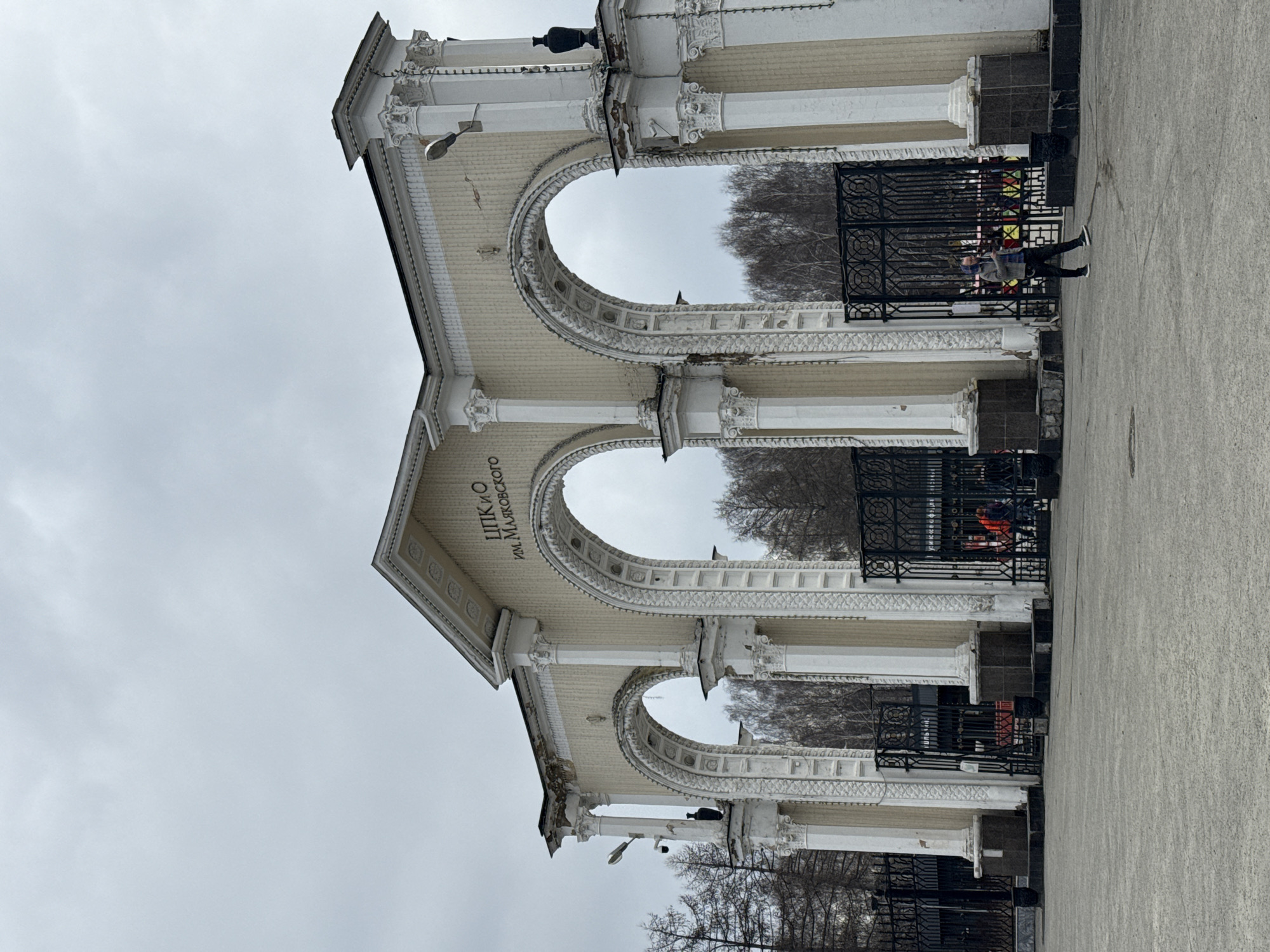 The image depicts the grand main entrance archway to the Central Park of Culture and Leisure named after V. Mayakovsky (ЦПКиО им. Маяковского) in Yekaterinburg, Russia, under an overcast sky. The archway, a large neoclassical structure, features three primary arched passages flanked by columns with Corinthian capitals and ornate white decorative elements. The main structure is a cream or pale yellow color, with white trim and fluted columns. The central arch bears the park's name in Russian on a pediment. The structure shows signs of age and weathering, including areas of peeling paint and discoloration. Streetlights and what appear to be CCTV cameras are mounted on the columns. Dark, urn-shaped decorative elements top some of the columns.
The arched passages are secured by tall, black wrought-iron gates with geometric patterns, which are currently closed. Through the gates, bare deciduous trees are visible in the background, suggesting late autumn, winter, or early spring. Several individuals in the distance, behind the gates, can be seen, some wearing brightly colored jackets (e.g., orange, yellow).
In the foreground, on a wide, grey concrete or asphalt plaza, a single individual, identified as male, walks from right to left. He wears a grey hooded jacket over a plaid shirt, dark trousers, and white-soled sneakers, and appears to be holding a small object or phone in his right hand. The ground surface is generally clean with some minor scuff marks.