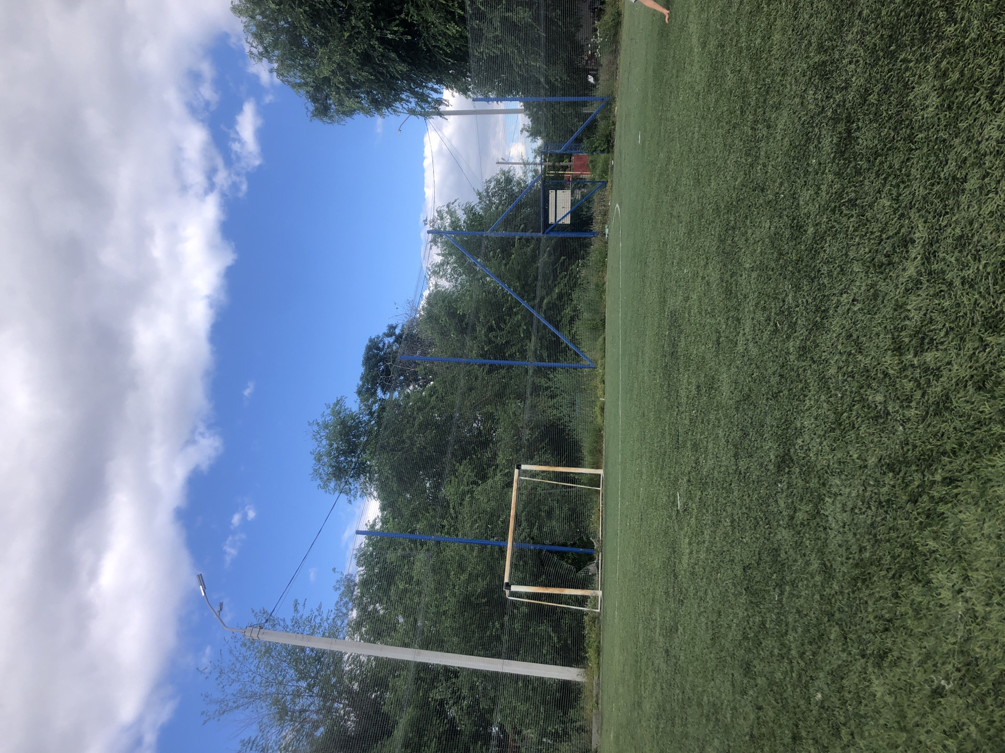 The image captures an outdoor artificial turf soccer field on a partly cloudy day. In the foreground, the green turf covers the majority of the frame. A goalpost, appearing weathered with some rust, stands prominently on the left side of the field. In the mid-ground, a fence made of netting with blue poles encloses the field. Trees and foliage surround the perimeter of the field, with a street light visible on the left. The background reveals a blue sky with scattered white and gray clouds. A portion of a person's arm and hand is visible on the far right side of the image, suggesting they are in motion or interacting with the field.